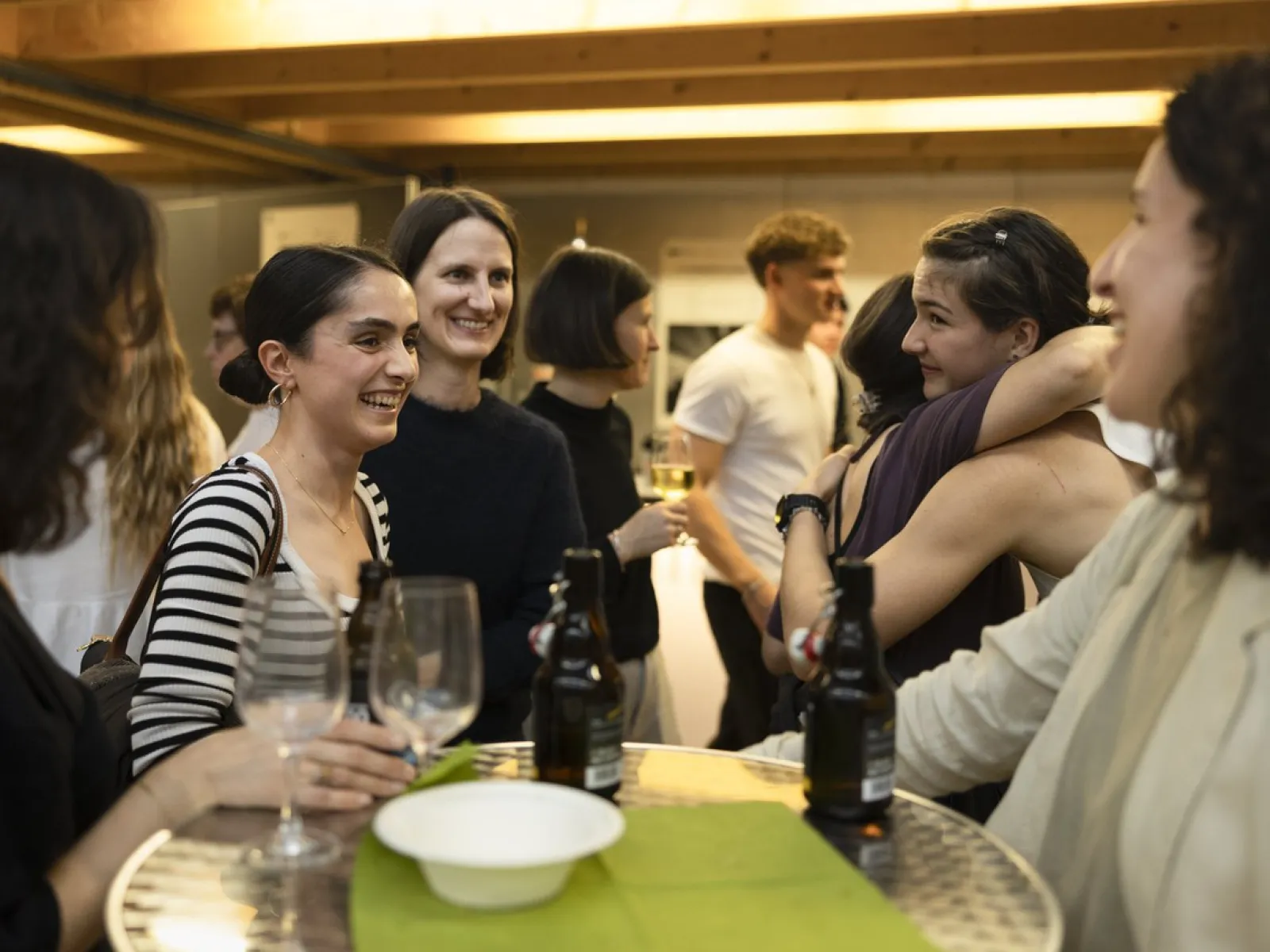 Eine Gruppe von Menschen steht um einen Tisch und unterhält sich fröhlich bei einem Event. Einige Personen halten Getränke in der Hand, während andere sich umarmen. Die Atmosphäre wirkt lebhaft und gesellig.