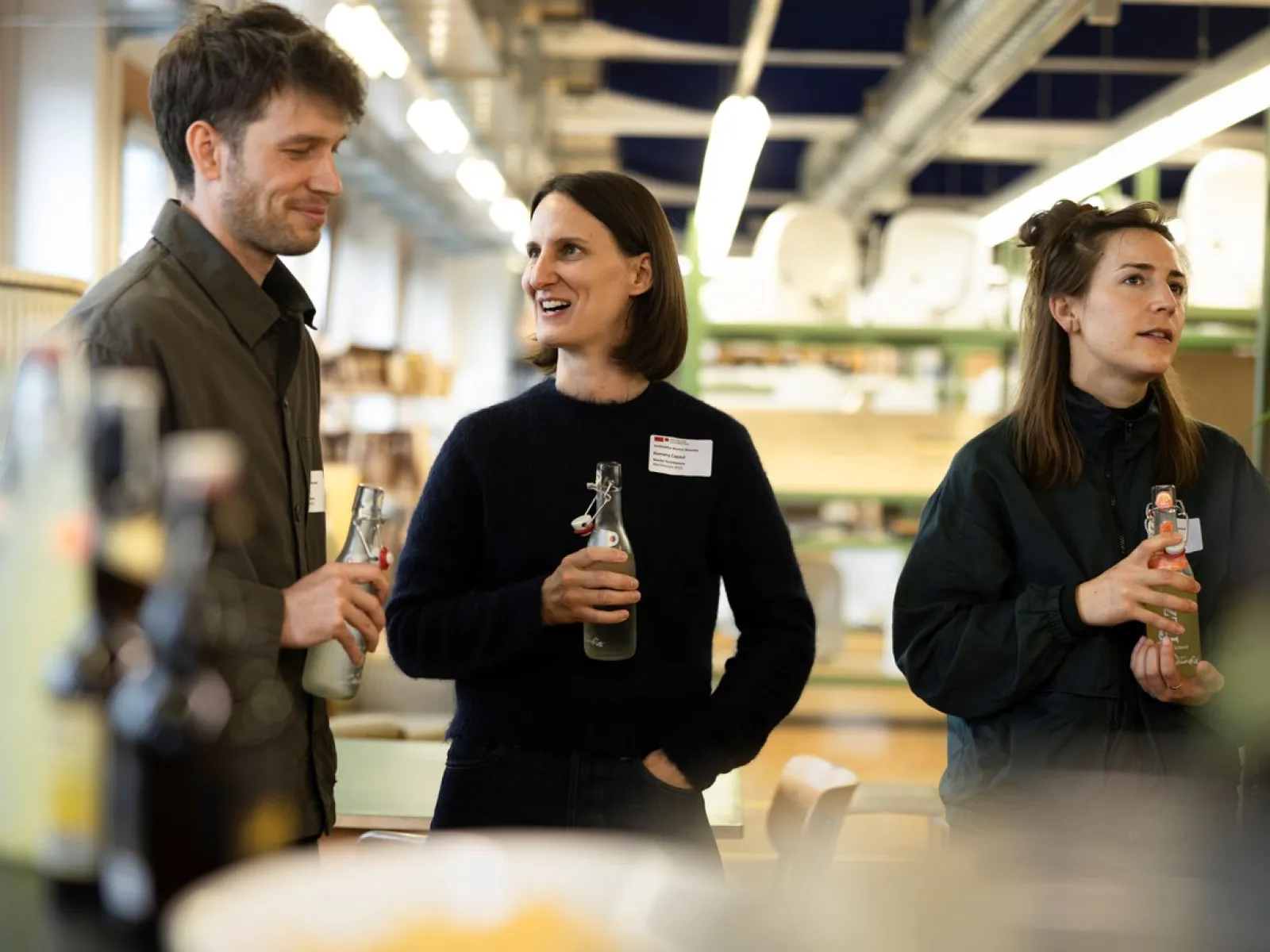Eine Gruppe von drei Personen steht in einem Innenraum und unterhält sich fröhlich. Zwei von ihnen halten Getränkeflaschen. Im Hintergrund sind Regale und Beleuchtung zu sehen. Die Atmosphäre wirkt lebhaft und gesellig.