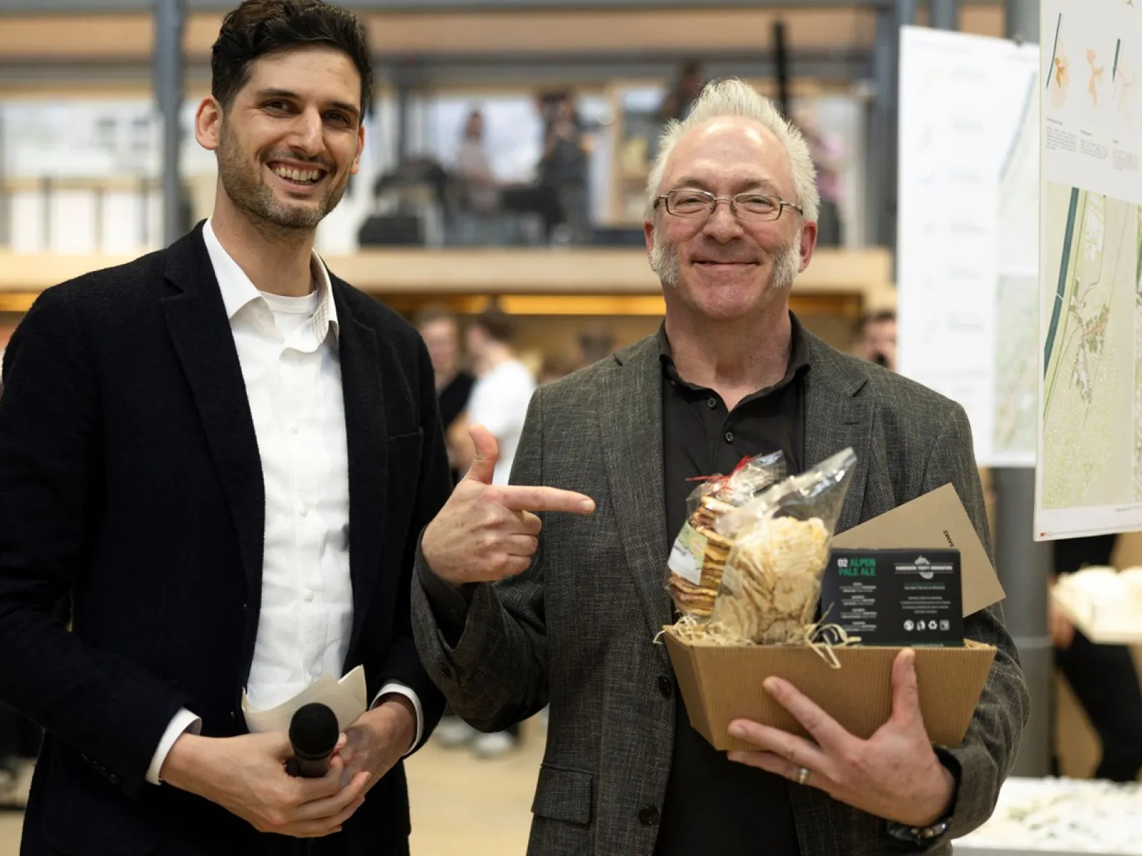 Zwei Männer stehen lächelnd in einem Innenraum. Der Mann links hält ein Mikrofon, während der Mann rechts auf einen Geschenkkorb zeigt, den er in der Hand hält. Im Hintergrund sind unscharf weitere Personen und eine moderne Architektur zu erkennen.