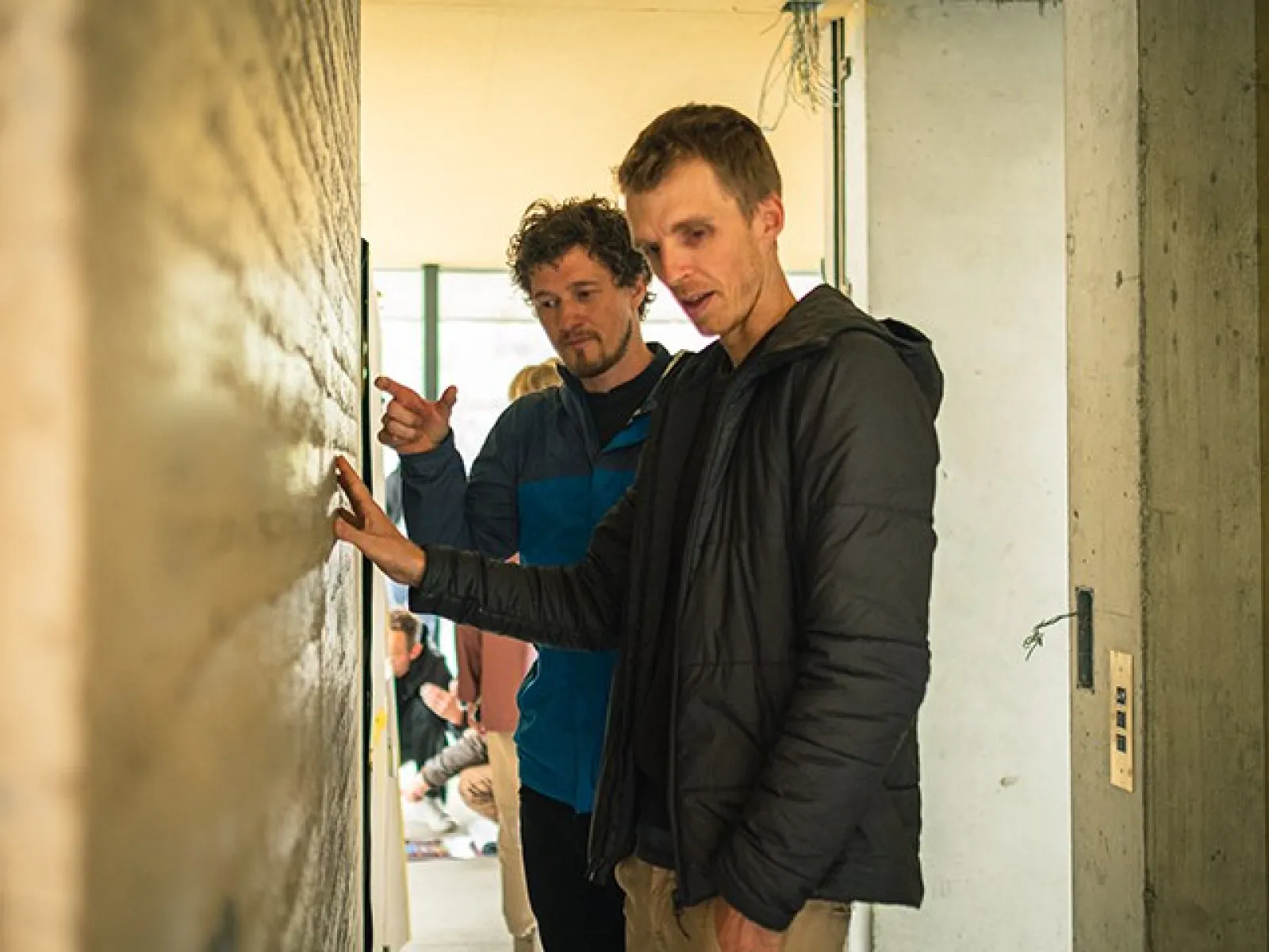 Zwei Männer betrachten und diskutieren eine Wand aus Stampflehm in einem Gebäude. Sie tragen Freizeitkleidung und befinden sich in einem hellen Raum.