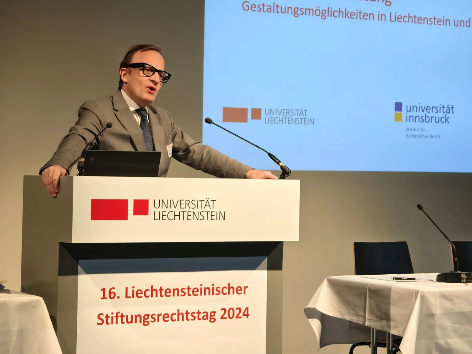 Ein Redner steht am Podium des 16. Liechtensteinischen Stiftungsrechtstags 2024 an der Universität Liechtenstein. Im Hintergrund sind Logos der Universität Liechtenstein und der Universität Innsbruck zu sehen.