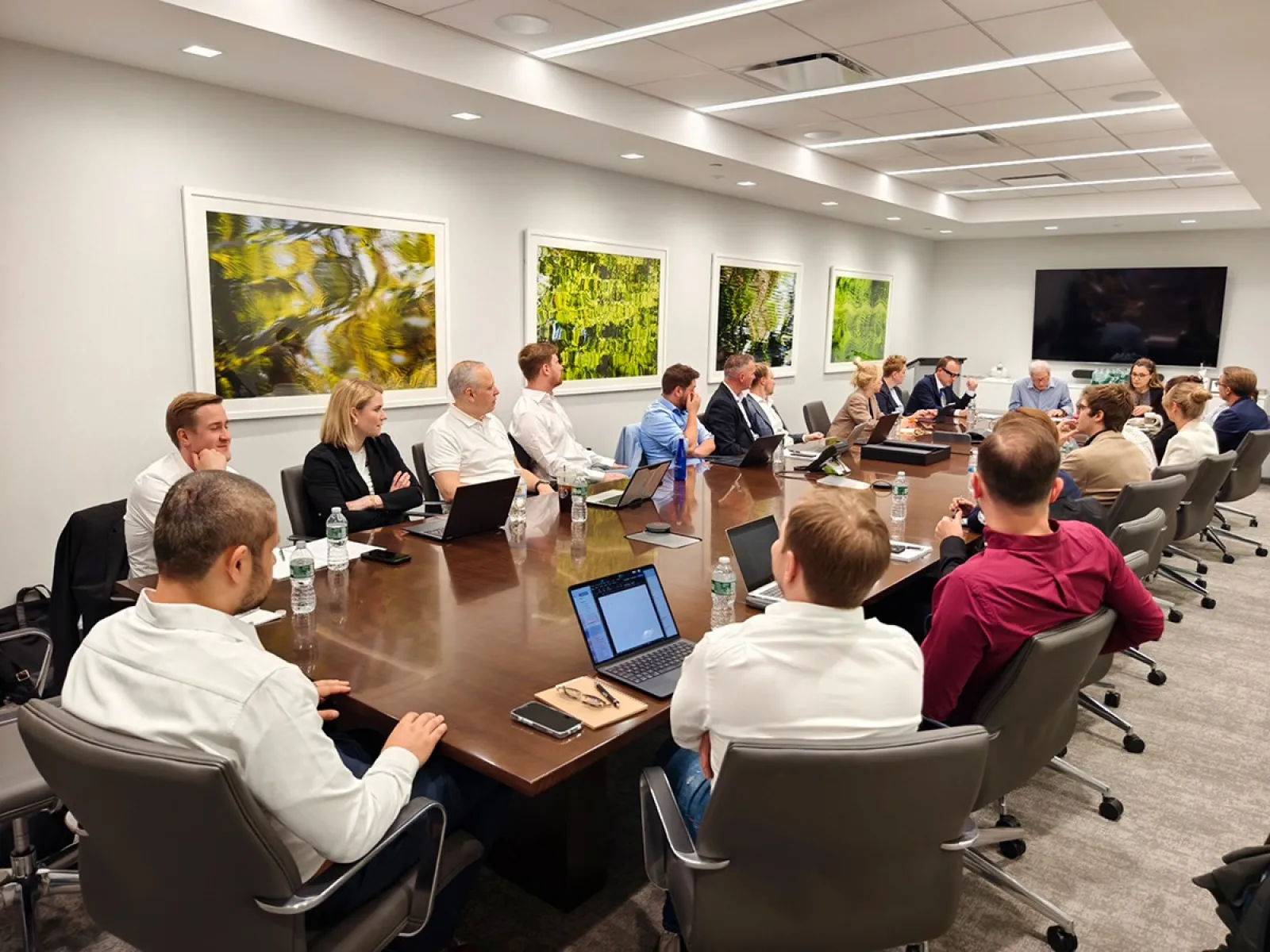 Eine Gruppe von Studierenden und Dozenten sitzt in einem modernen Konferenzraum an einem großen Tisch. Sie sind in eine Diskussion vertieft, umgeben von Laptops und Notizen. An den Wänden hängen Kunstwerke, und im Hintergrund ist ein großer Bildschirm zu sehen.