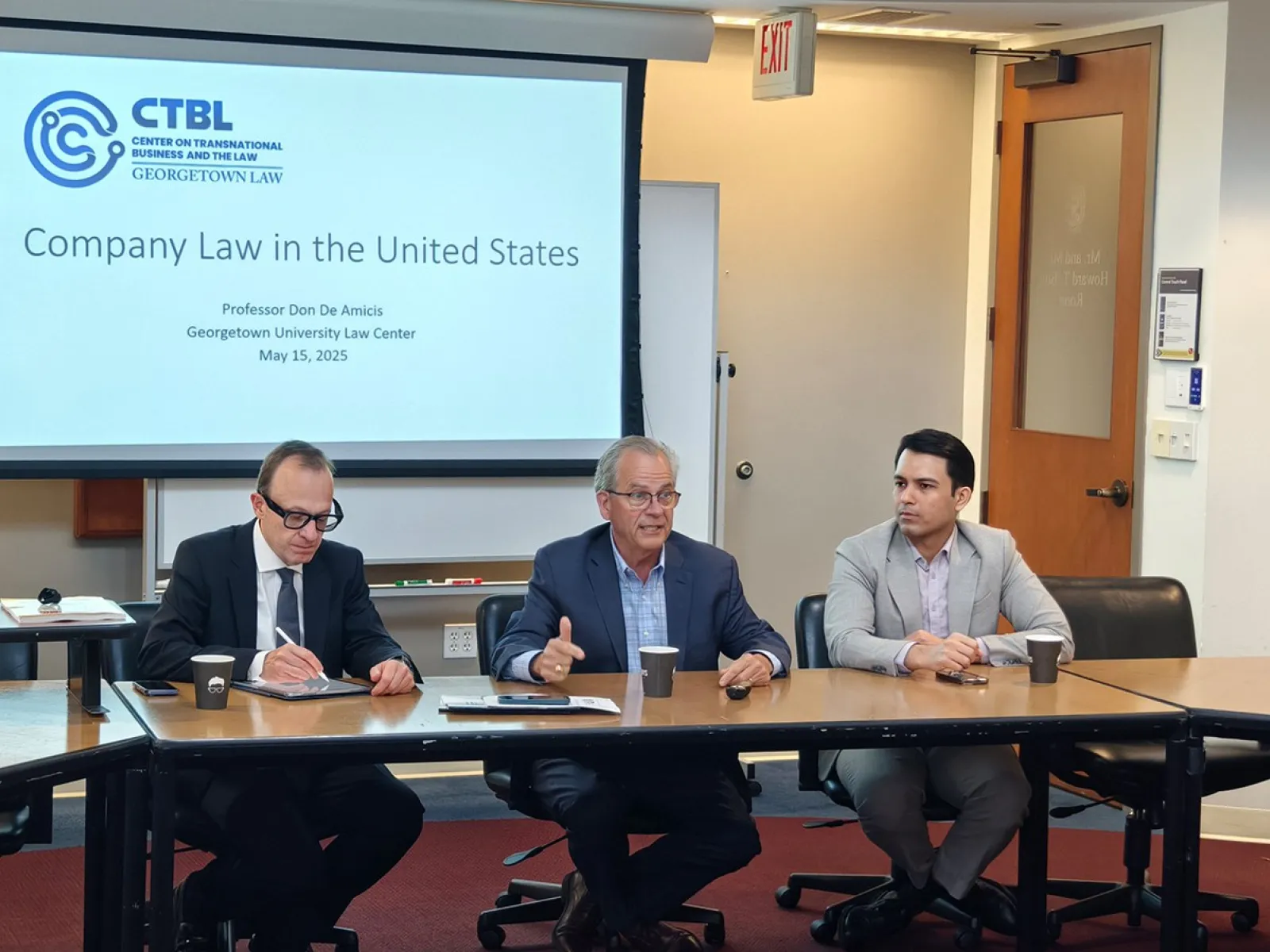 Drei Personen sitzen an einem Konferenztisch in einem Seminarraum. Im Hintergrund ist eine Präsentationsfolie mit dem Titel "Company Law in the United States" zu sehen. Der Raum gehört zum Georgetown University Law Center.