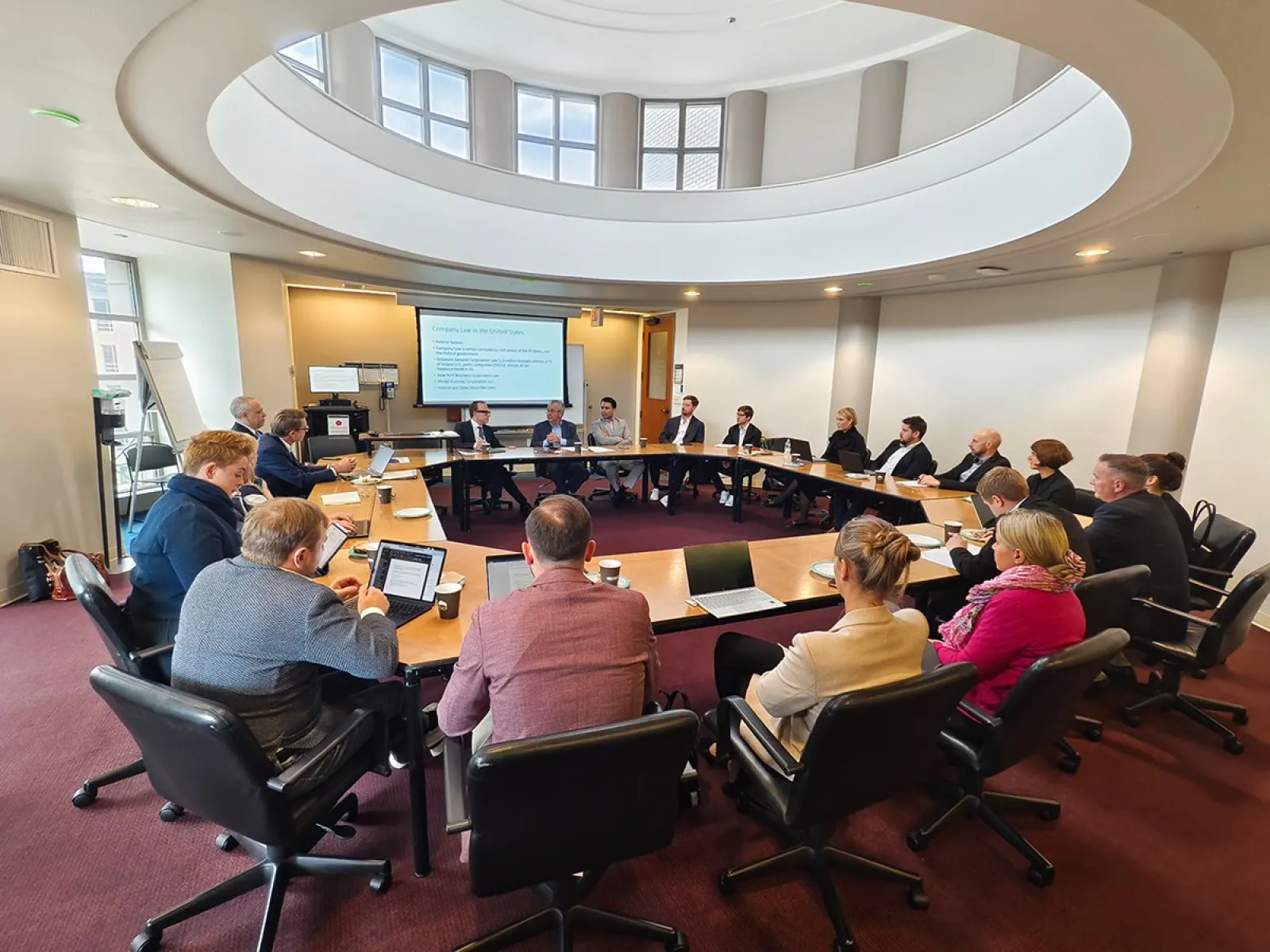Teilnehmende einer Studienreise sitzen in einem modernen Konferenzraum an einem ovalen Tisch. Eine Präsentation wird auf einem Bildschirm gezeigt.