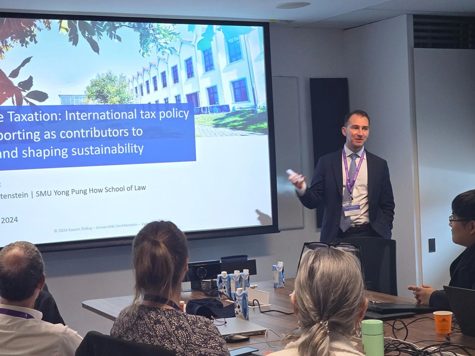 Ein Vortragender steht vor einer Präsentation mit dem Titel "Sustainable Taxation: International tax policy and reporting as contributors to achieving and shaping sustainability". Im Raum sitzen mehrere Personen, die dem Vortrag folgen.