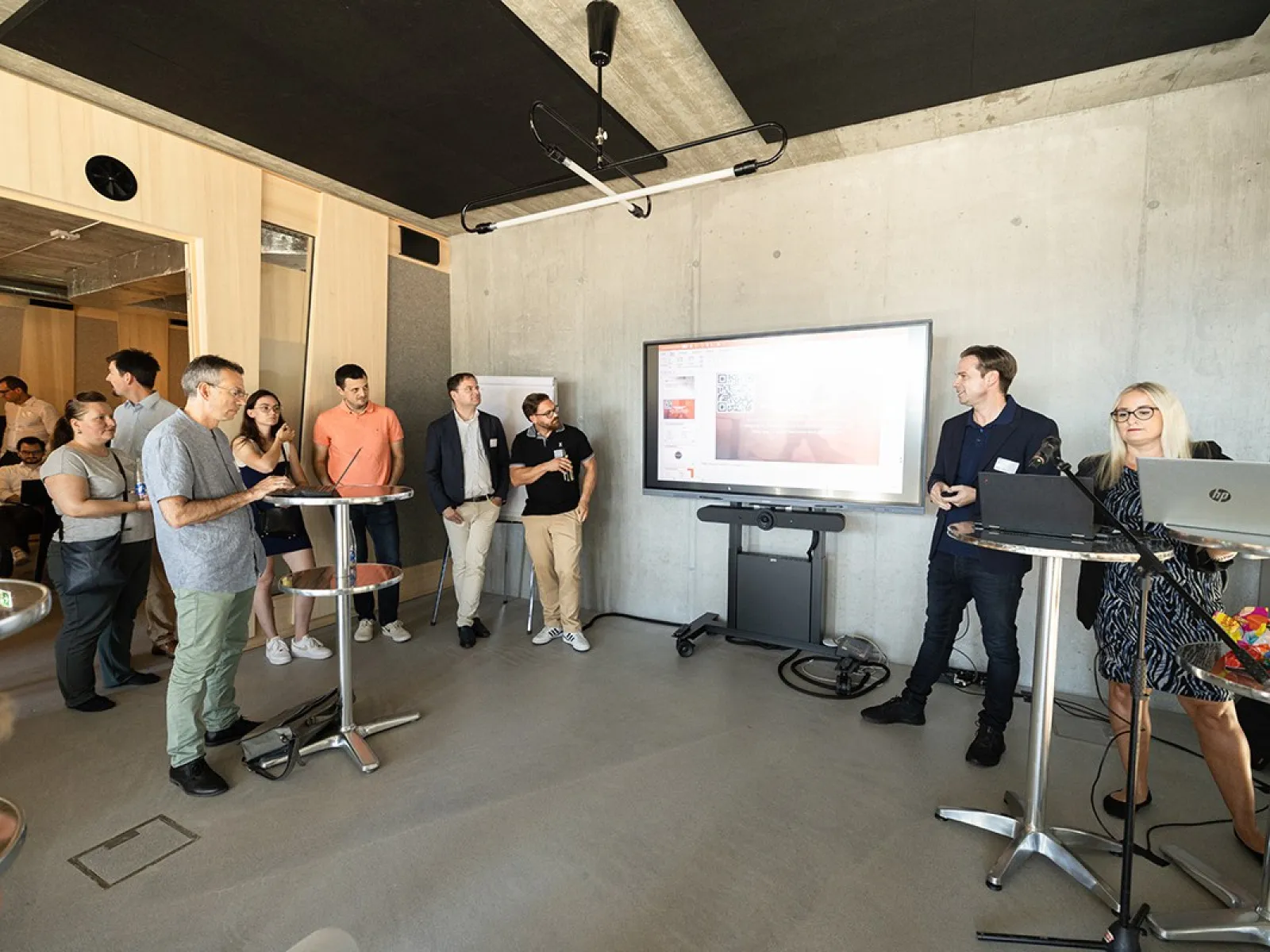 Eine Gruppe von Menschen steht in einem modernen Raum mit Betonwänden. Im Vordergrund sind Stehtische und ein großer Bildschirm an der Wand, auf dem eine Präsentation gezeigt wird. Eine Person spricht zu den Anwesenden, während andere aufmerksam zuhören. Der Raum wirkt hell und einladend.