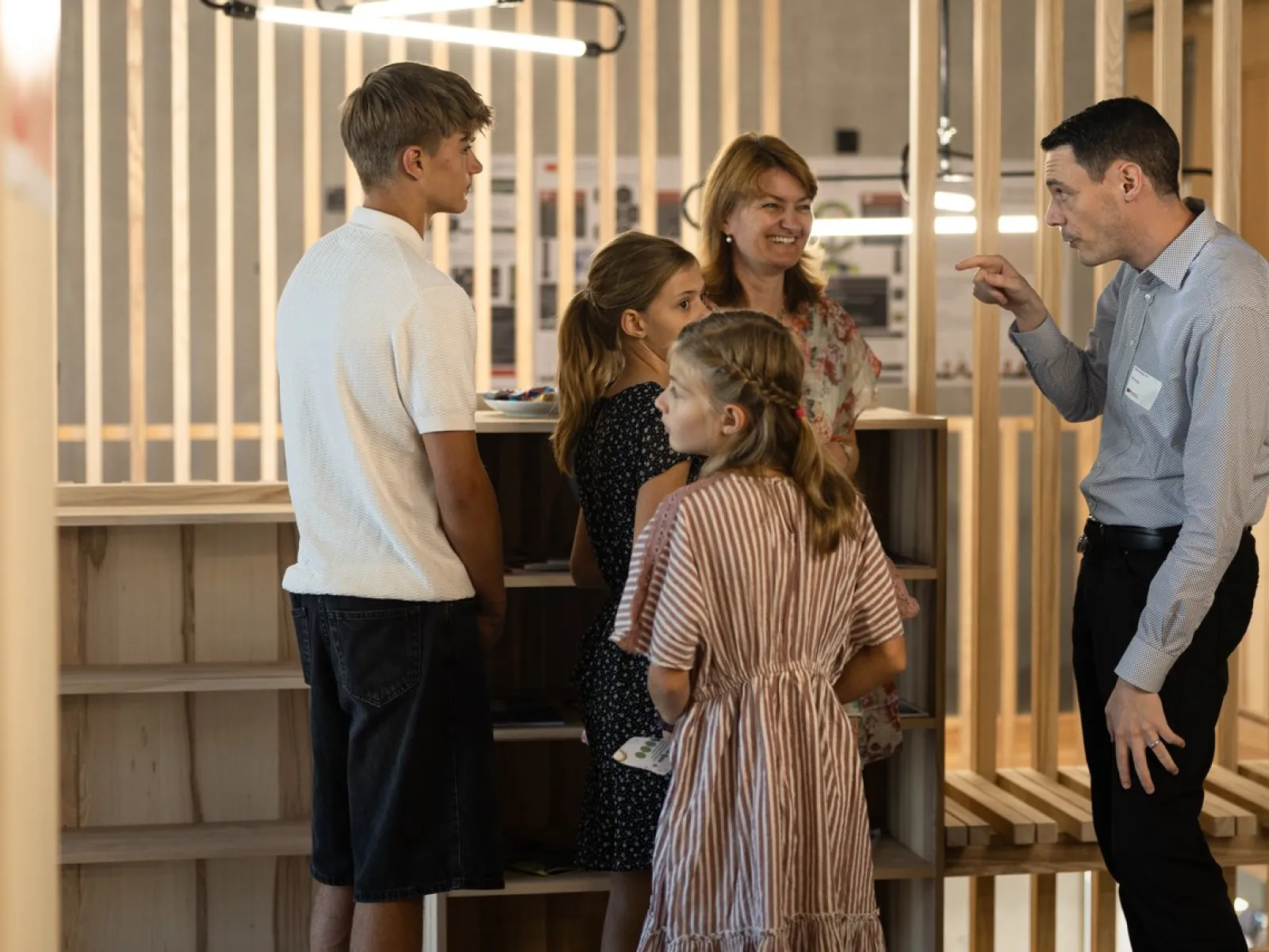 Eine Gruppe von Menschen steht in einem modernen Raum mit Holzelementen. Ein Mann spricht gestikulierend mit einer Frau und drei Kindern. Die Szene vermittelt eine freundliche und einladende Atmosphäre während des Tags der offenen Tür an der Universität Liechtenstein.
