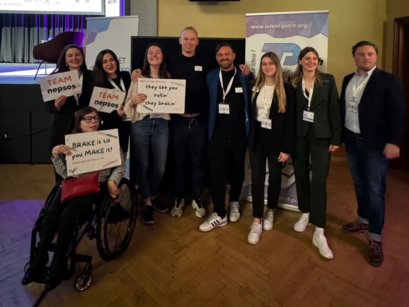 Gruppe von neun Personen posiert lächelnd bei einem Event, einige halten Schilder mit motivierenden Botschaften. Im Hintergrund sind Banner mit der Aufschrift "Talent Pitch" zu sehen. Eine Person sitzt im Rollstuhl.