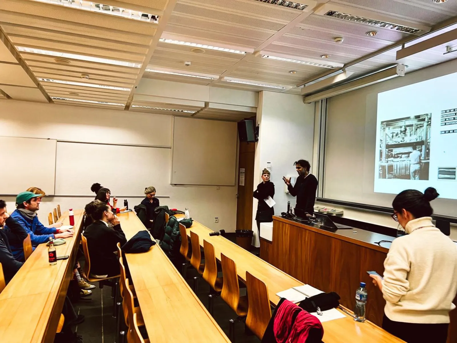 Ein Vortrag in einem Hörsaal: Zwei Personen präsentieren vor einer Gruppe von Studierenden. Auf der Leinwand ist eine Projektion zu sehen. Die Studierenden sitzen an Holztischen und hören aufmerksam zu.