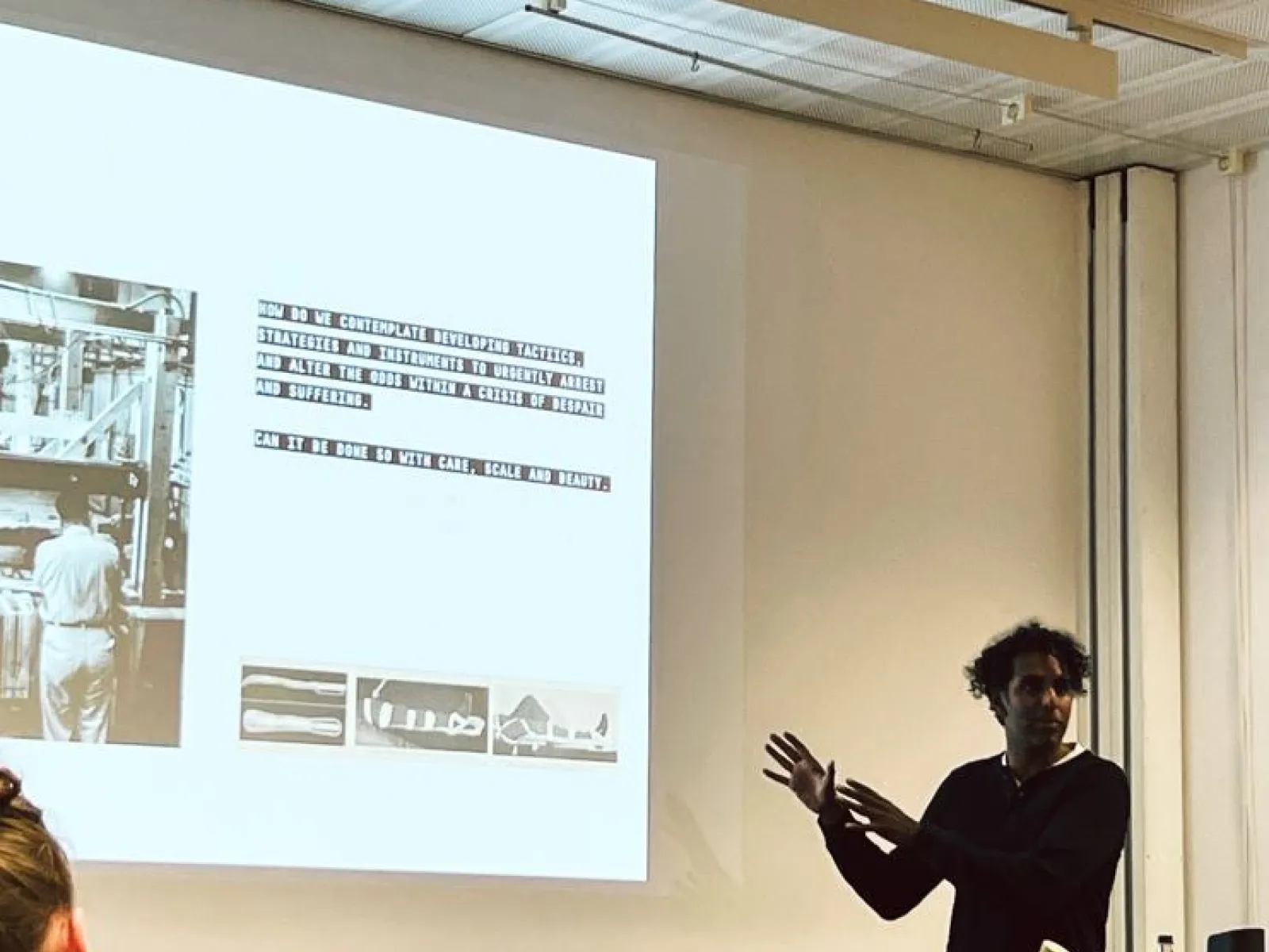 Ein Vortragender steht vor einer Projektionsleinwand und präsentiert Inhalte zu urbanem Design. Auf der Leinwand sind Text und Bilder zu sehen. Im Vordergrund sitzen Zuhörer. Der Raum wirkt modern und technisch ausgestattet.