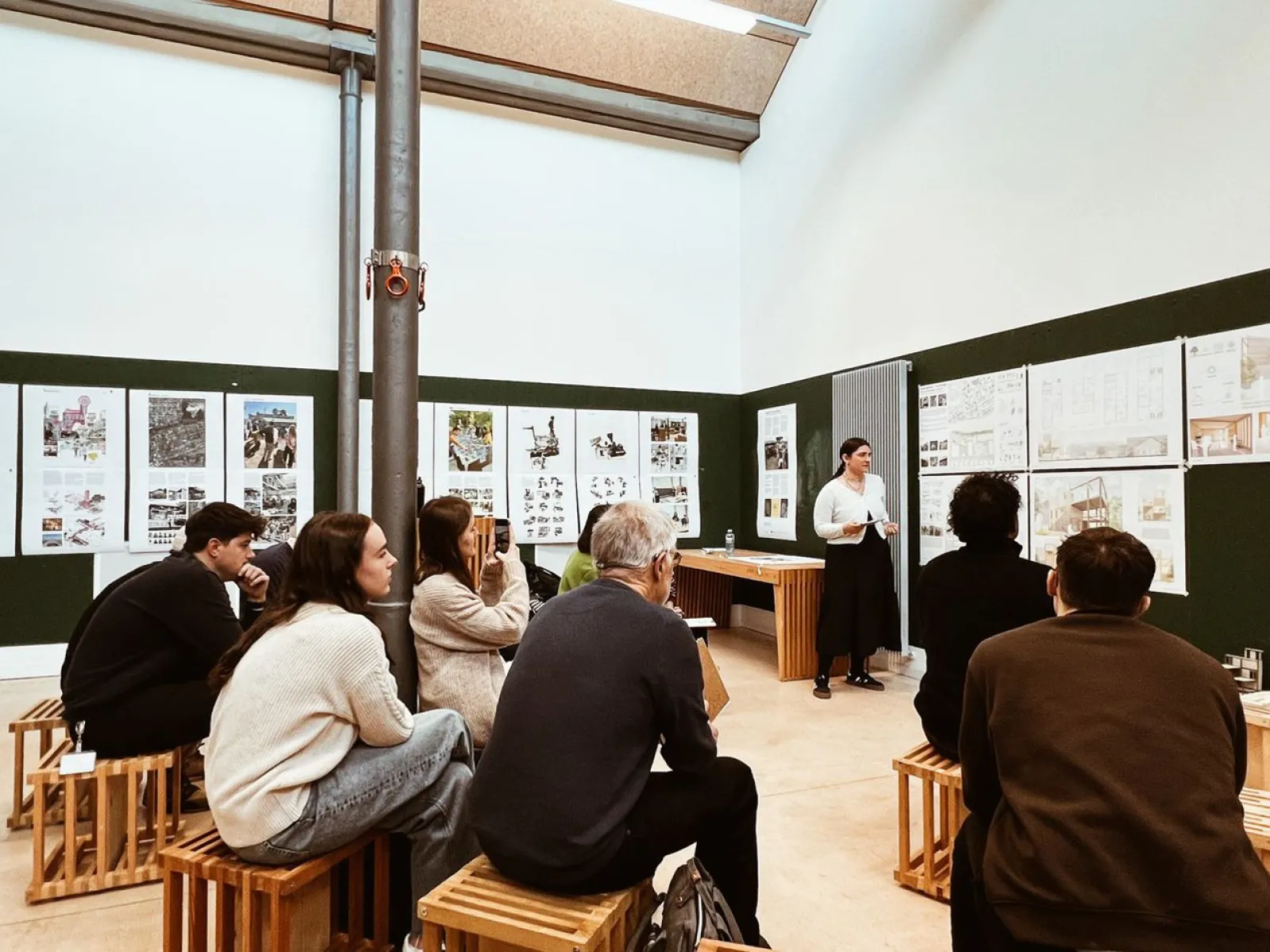 Eine Gruppe von Studierenden sitzt auf Holzhockern in einem Seminarraum und hört einer Person zu, die vor Präsentationspostern an der Wand steht. Die Atmosphäre ist konzentriert und akademisch.