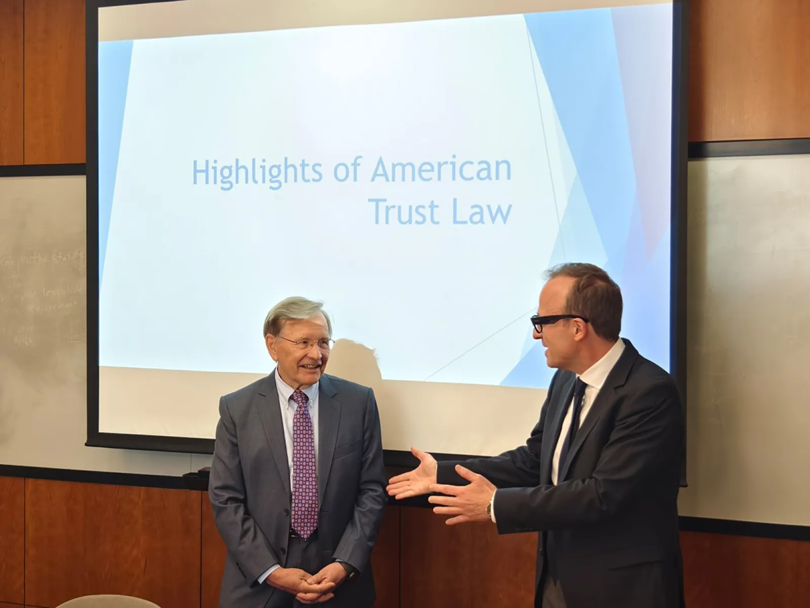 Zwei Männer in Anzügen stehen vor einer Präsentation mit dem Titel "Highlights of American Trust Law". Einer der Männer spricht gestikulierend. Im Hintergrund ist ein Projektionsbildschirm zu sehen.