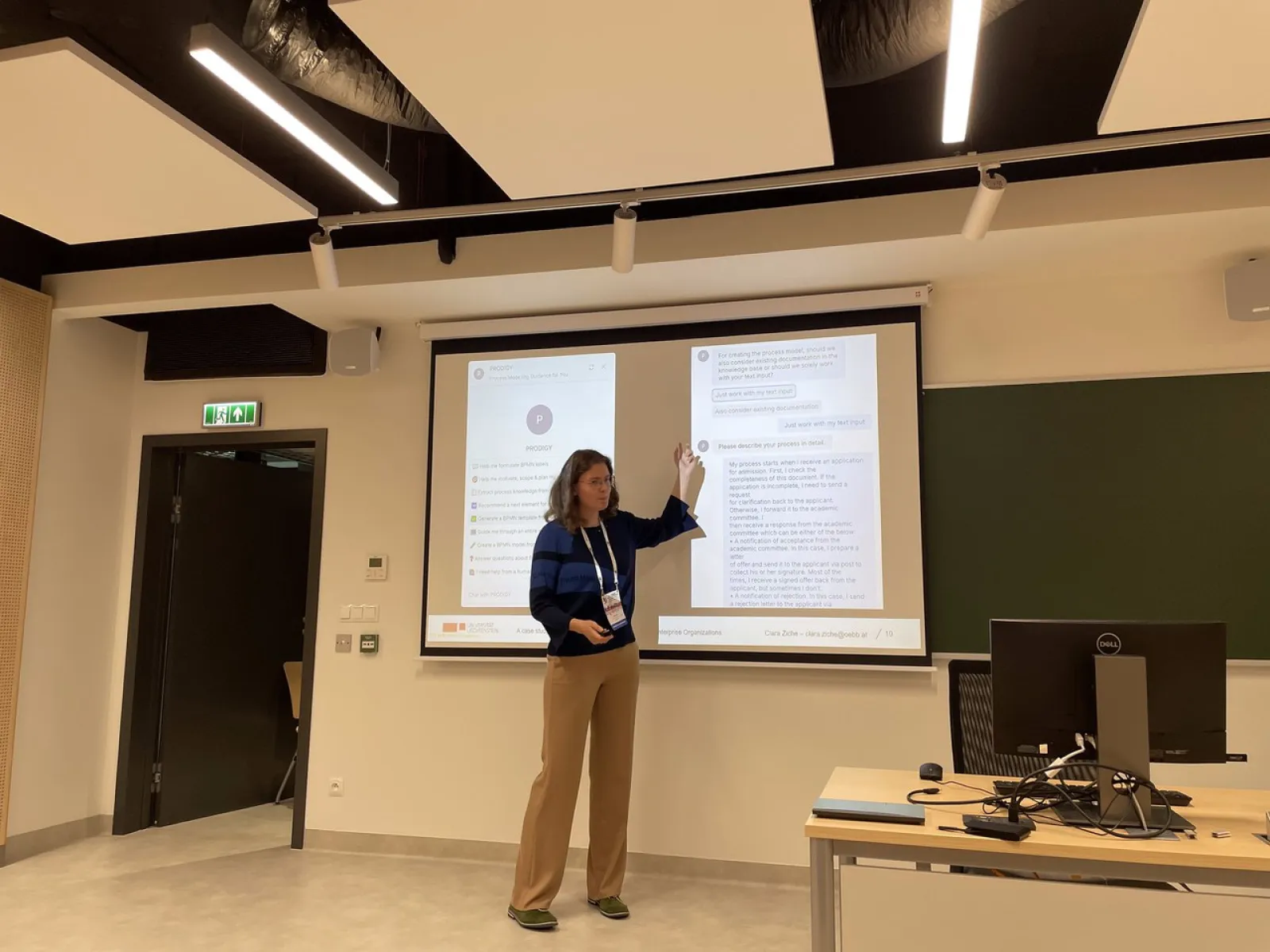 Eine Person präsentiert in einem modernen Seminarraum vor einer Leinwand mit zwei projizierten Dokumenten. Im Vordergrund steht ein Tisch mit einem Computerbildschirm.