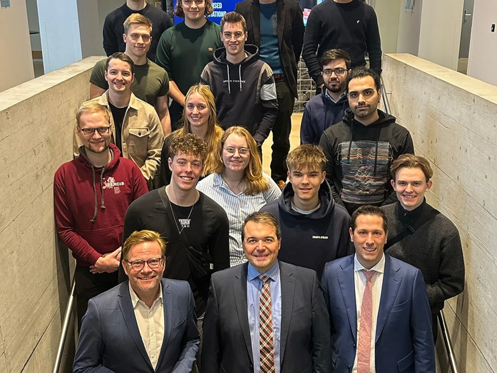 Gruppe von Studierenden und Lehrenden steht auf einer Treppe in einem modernen Gebäude der Universität Liechtenstein, lächelnd in die Kamera. Im Hintergrund sind Bildschirme und eine Steinwand zu sehen.