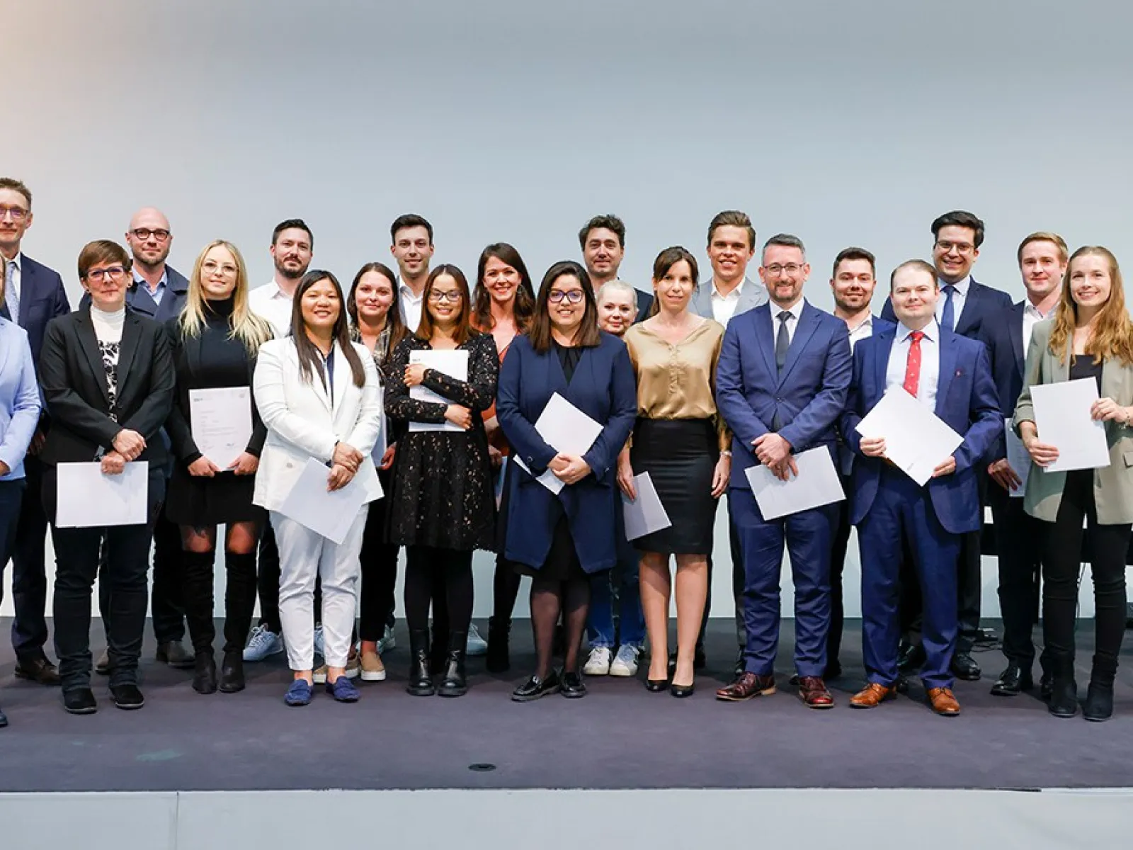 Eine Gruppe von Absolventinnen und Absolventen steht auf einer Bühne und hält Zertifikate in den Händen. Sie lächeln in die Kamera. Die Personen sind formell gekleidet, und im Hintergrund ist eine neutrale Wand zu sehen.