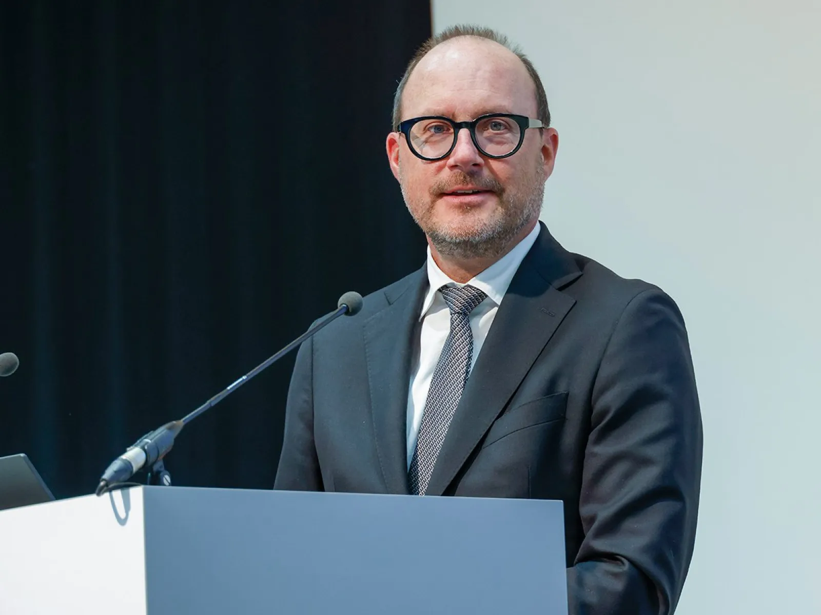 Ein Mann in Anzug und Krawatte steht an einem Rednerpult und spricht in ein Mikrofon. Der Hintergrund ist schlicht und professionell gestaltet.