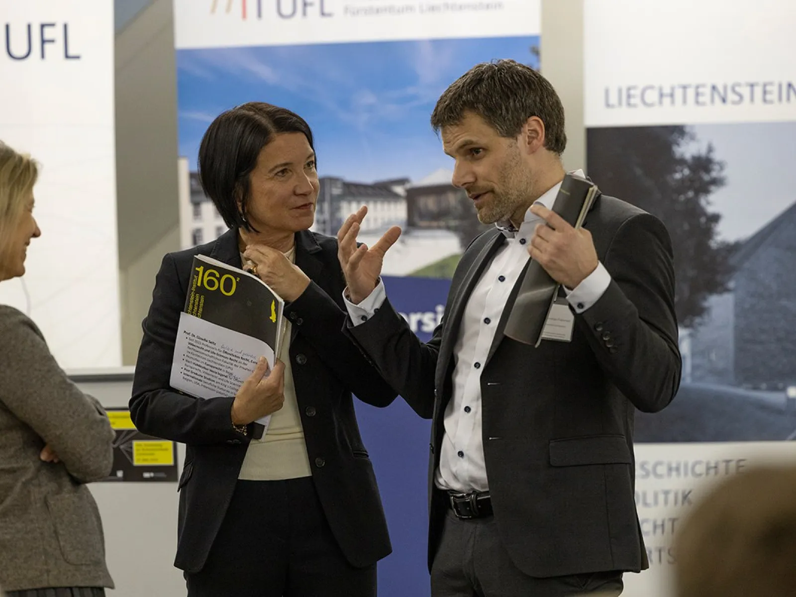 Drei Personen stehen in einem Gespräch vor Roll-Up-Bannern der Universität Liechtenstein und des Liechtenstein-Instituts. Eine Person hält ein Buch in der Hand. Die Szene findet in einem formellen Rahmen statt, vermutlich bei einer akademischen Veranstaltung.