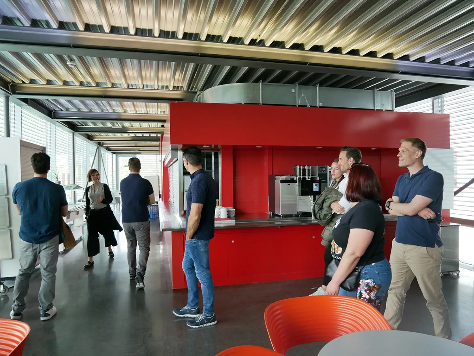 Eine Gruppe von Menschen steht in einem modernen Raum mit roten Wänden und einer Kaffeestation, möglicherweise während eines Treffens an der Universität Liechtenstein.