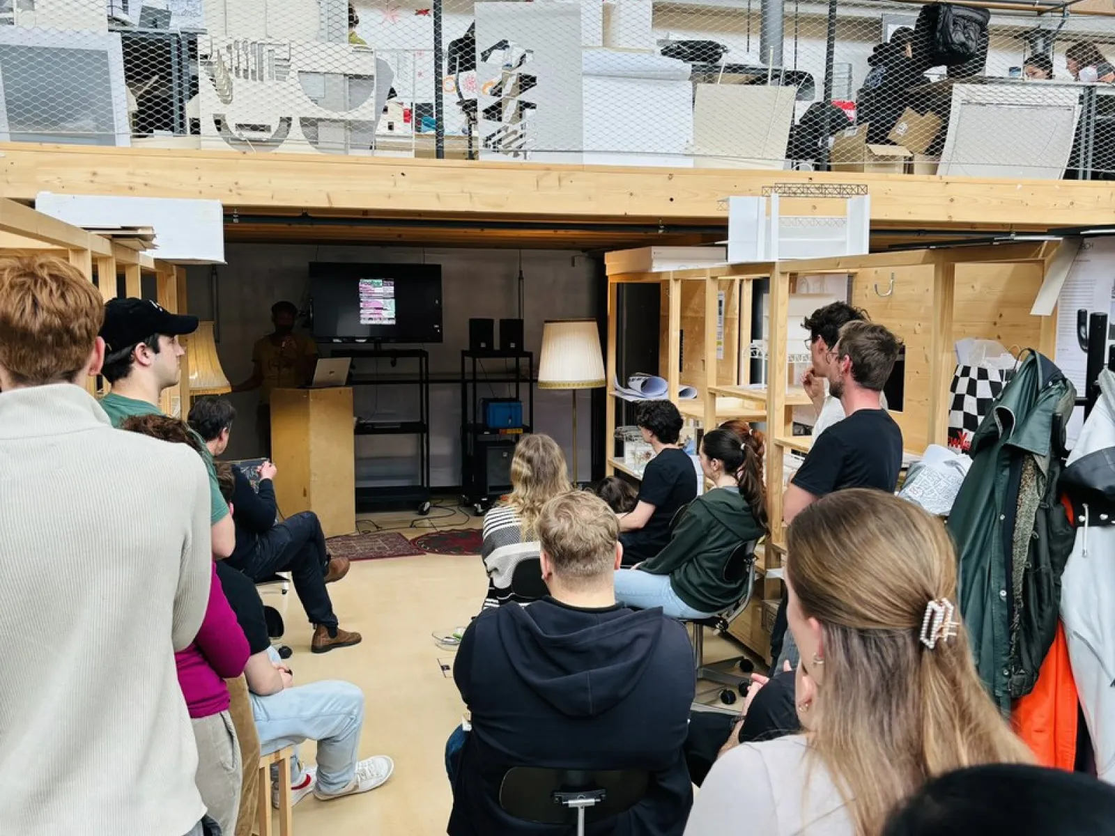Eine Gruppe von Studierenden sitzt in einem Architekturatelier und hört einem Vortrag zu. Im Hintergrund sind Regale und Arbeitsmaterialien zu sehen.