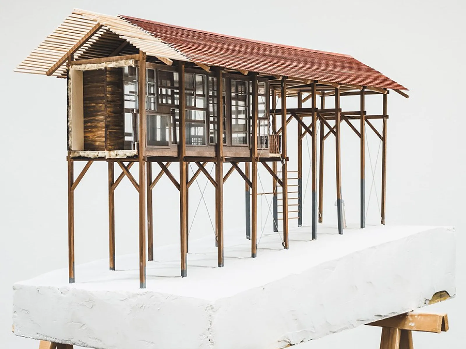 Ein Architekturmodell eines Strickbaus auf Stelzen, das auf einem weißen Sockel präsentiert wird. Das Modell zeigt eine lineare Struktur mit einem Satteldach und großen Fenstern, die traditionelle Bauweise und moderne Elemente kombinieren.