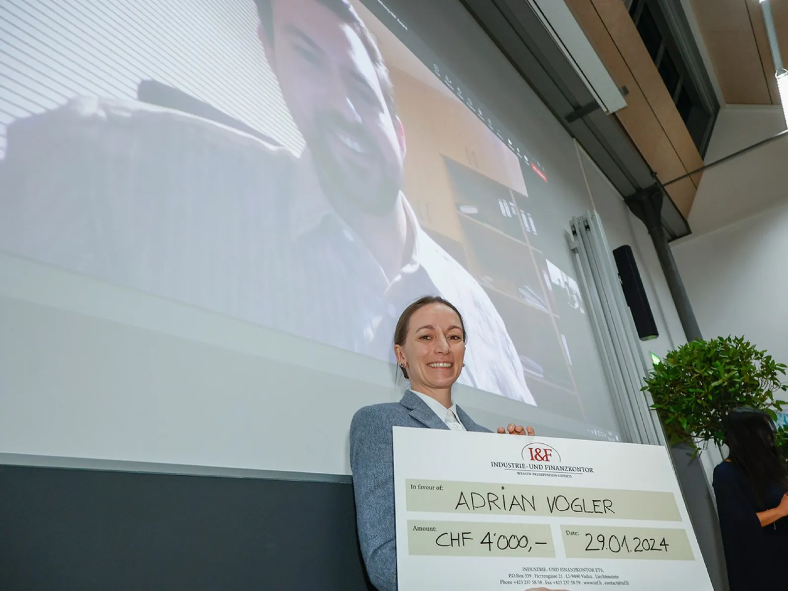 Eine Person hält einen großen Scheck mit der Aufschrift "Adrian Vogler, CHF 4,000, 29.01.2024" in einem Vortragsraum. Im Hintergrund ist eine Projektion eines lächelnden Mannes auf einer Leinwand zu sehen.