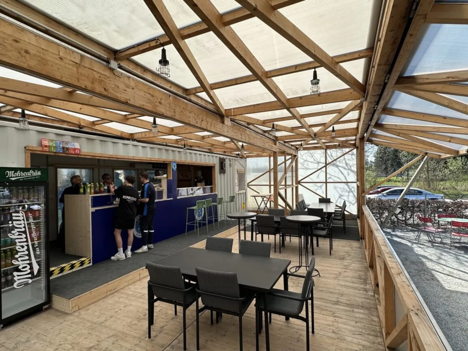 Ein moderner, überdachter Raum mit Holzkonstruktion und transparentem Dach. Links befindet sich eine Bar mit Personen, die bedient werden. Rechts stehen Tische und Stühle für Besucher bereit. Die Umgebung wirkt einladend und funktional für Veranstaltungen oder Treffen.