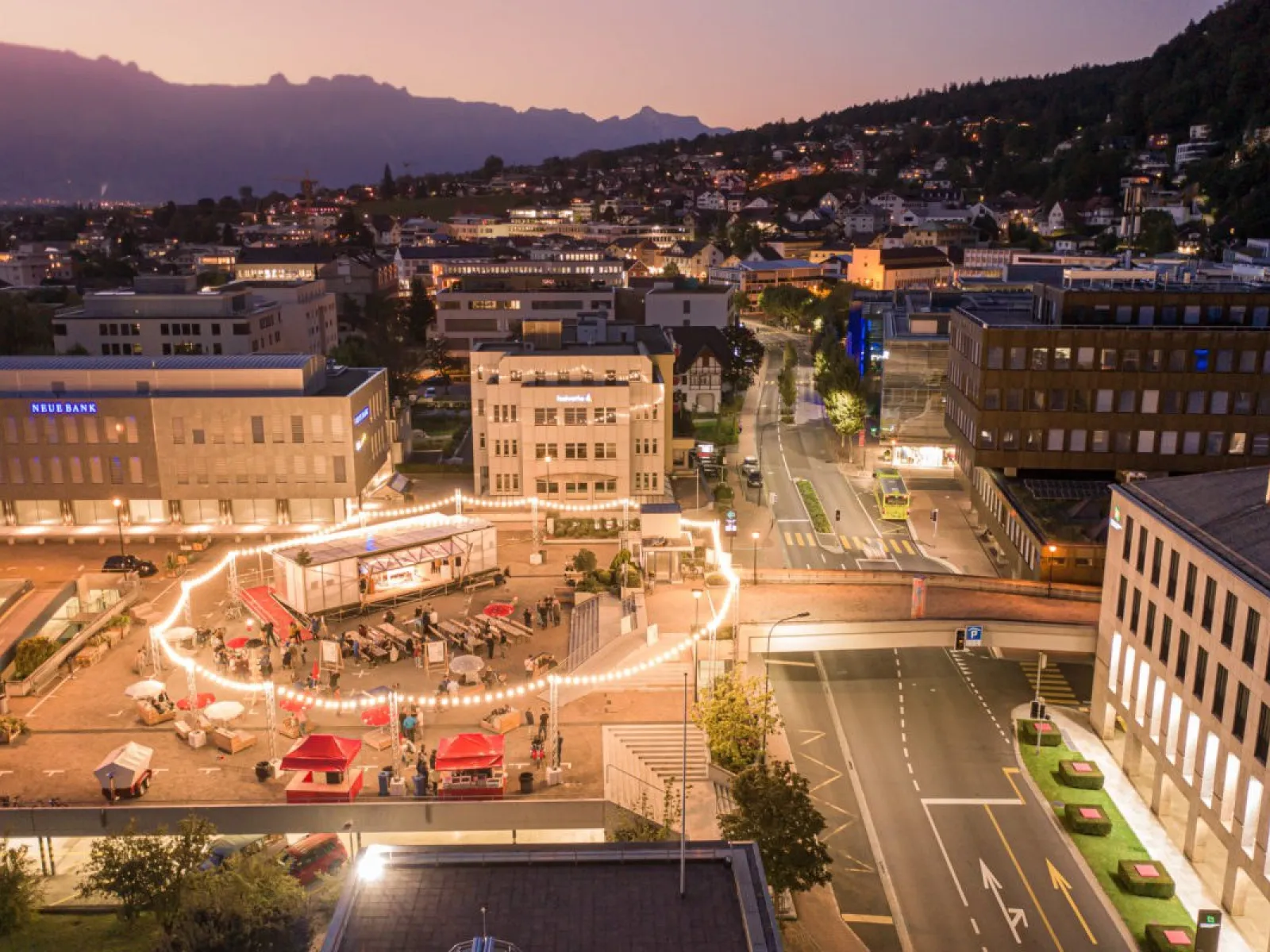 Luftaufnahme einer städtischen Umgebung bei Dämmerung. Im Zentrum des Bildes ist ein beleuchteter Platz mit einem kreisförmigen Lichterkranz zu sehen, umgeben von Gebäuden. Menschen versammeln sich um das Base Camp, eine modulare Plattform. Im Hintergrund erstreckt sich eine hügelige Landschaft.