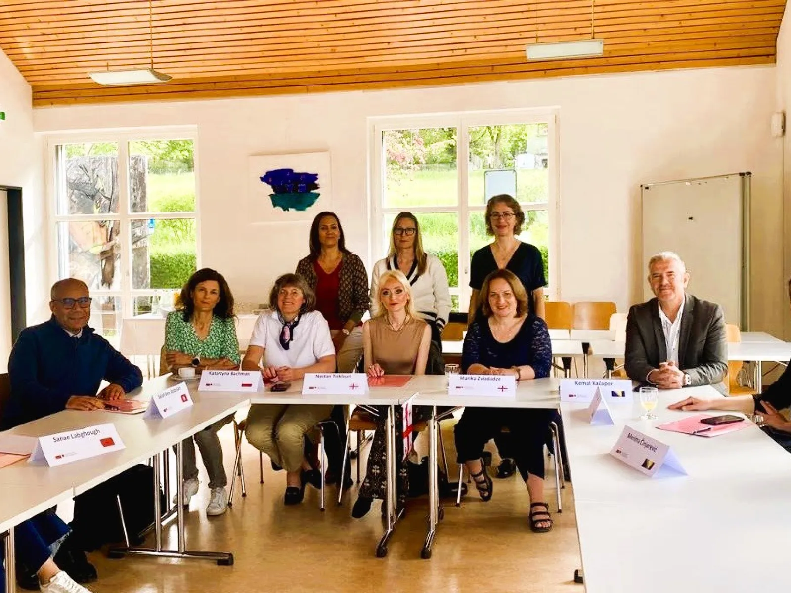 Eine Gruppe von zwölf Personen sitzt an einem U-förmigen Tisch in einem hellen Raum. Sie sind Teil eines internationalen Besuchsprogramms an der Universität Liechtenstein. Im Hintergrund sind große Fenster und eine Holzbalkendecke zu sehen.
