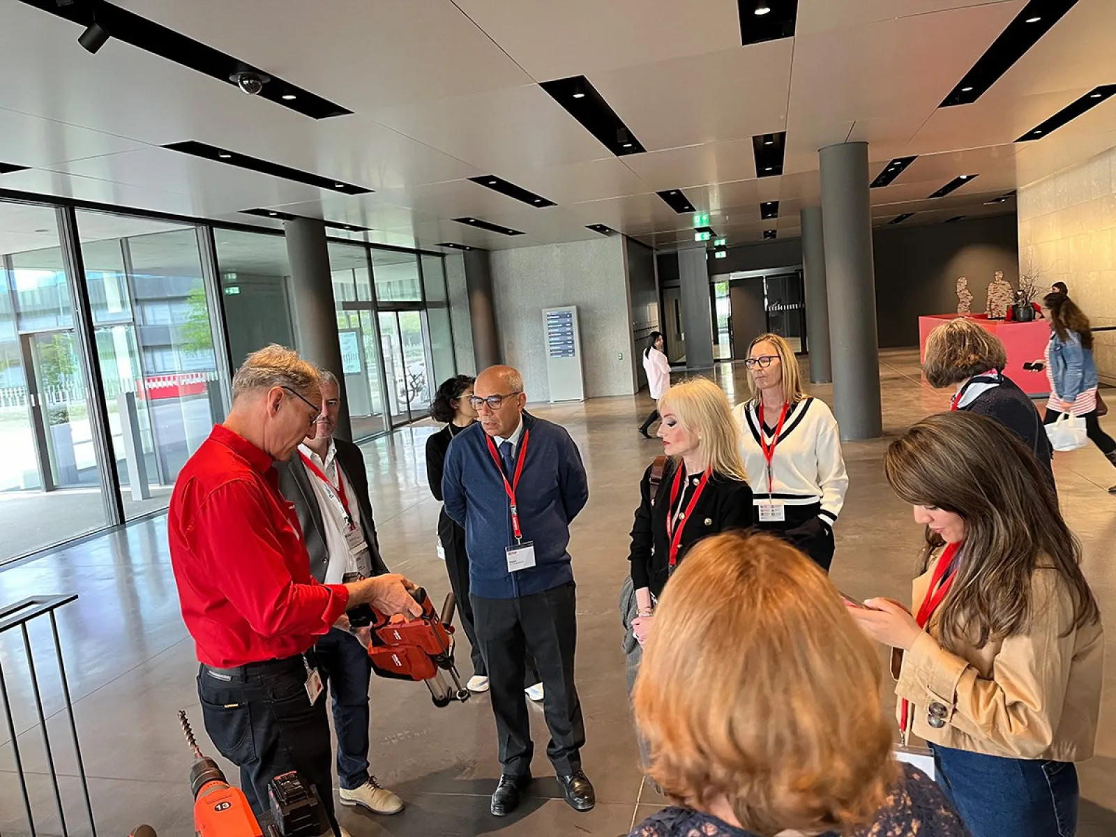 Eine Gruppe von Menschen steht in einem modernen Gebäude und hört einer Person zu, die etwas erklärt. Alle tragen Namensschilder und einige haben rote Lanyards. Im Hintergrund sind große Fenster und moderne Architektur zu sehen.