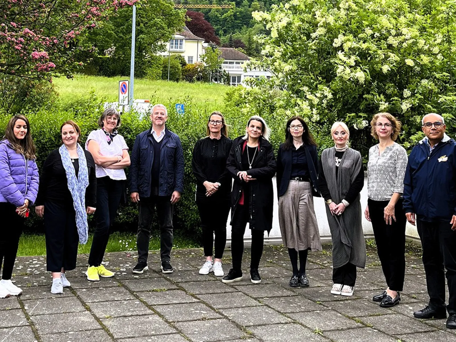 Eine Gruppe von zehn Personen steht draußen auf einem gepflasterten Weg vor einer grünen Hecke und blühenden Bäumen. Die Personen sind in Freizeitkleidung und lächeln in die Kamera. Im Hintergrund sind ein Gebäude und eine Wiese zu sehen.