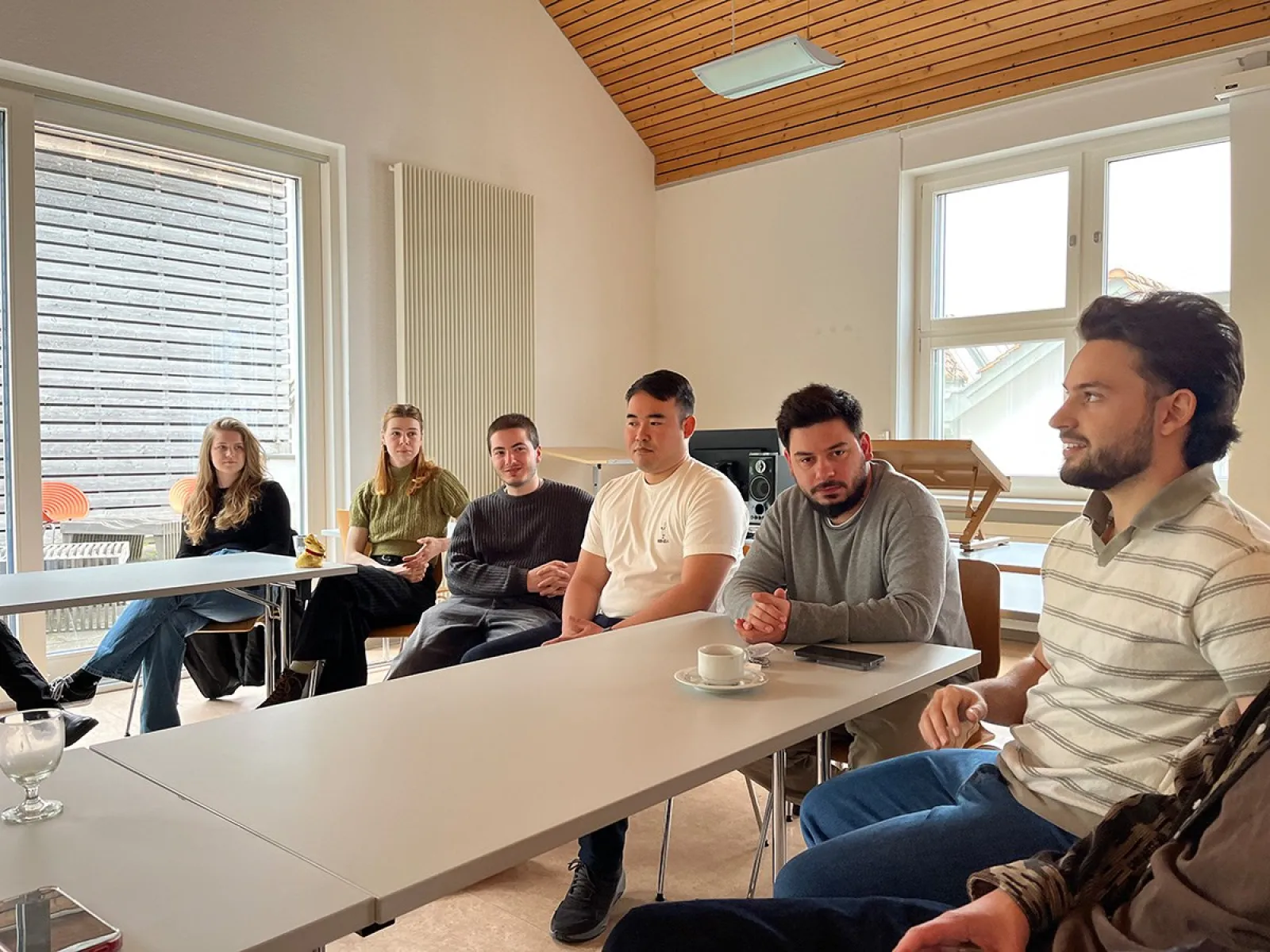 Eine Gruppe von Menschen sitzt in einem Besprechungsraum an Tischen. Sie sind in ein Gespräch vertieft. Im Hintergrund sind große Fenster und eine Holzdecke zu sehen.