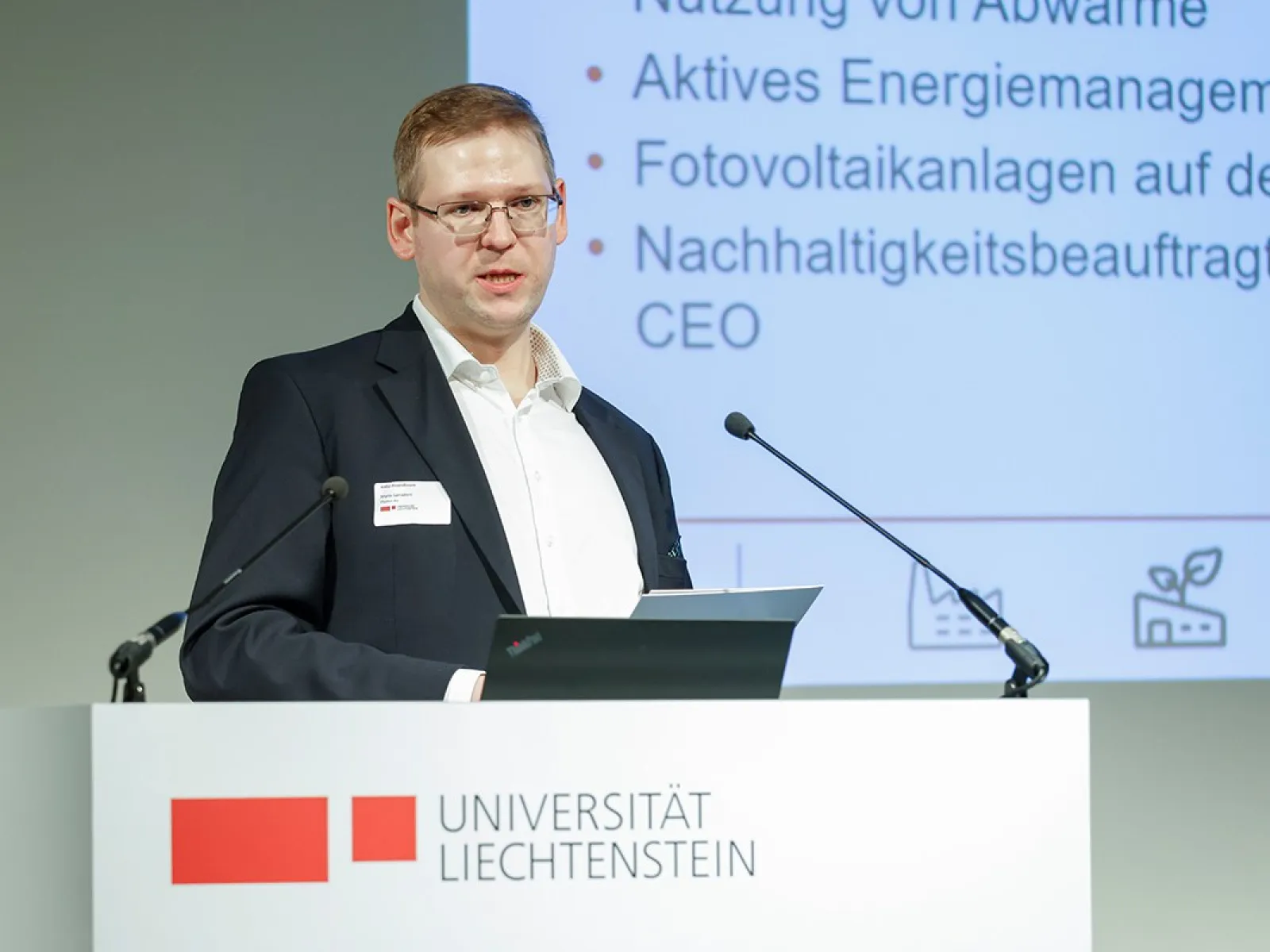 Ein Mann hält einen Vortrag an einem Rednerpult der Universität Liechtenstein. Im Hintergrund ist eine Präsentation mit Themen wie Energiemanagement und Photovoltaikanlagen zu sehen.