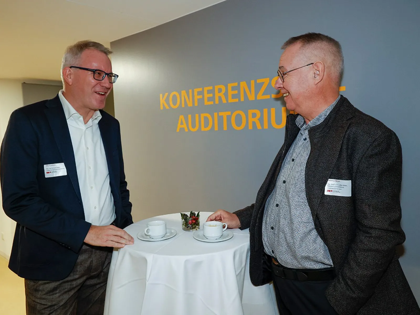 Zwei Männer im Gespräch bei einer Veranstaltung an der Universität Liechtenstein. Sie stehen an einem Stehtisch mit Kaffeetassen im Bereich "Konferenzzentrum Auditorium".