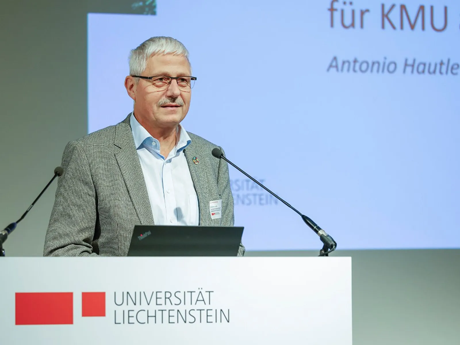 Ein Redner steht an einem Podium mit dem Logo der Universität Liechtenstein und spricht in ein Mikrofon. Im Hintergrund ist eine Präsentationsfolie zu sehen.