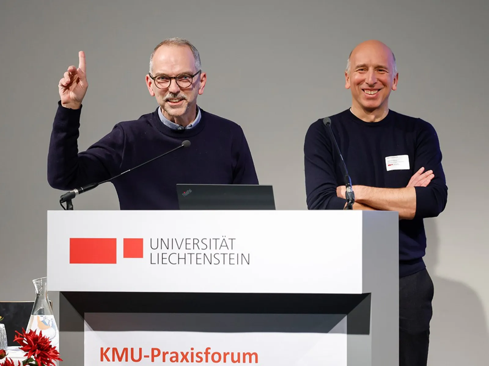 Zwei Männer stehen lächelnd hinter einem Rednerpult mit dem Logo der Universität Liechtenstein. Einer hebt die Hand, während der andere die Arme verschränkt. Im Vordergrund sind rote Blumen zu sehen.