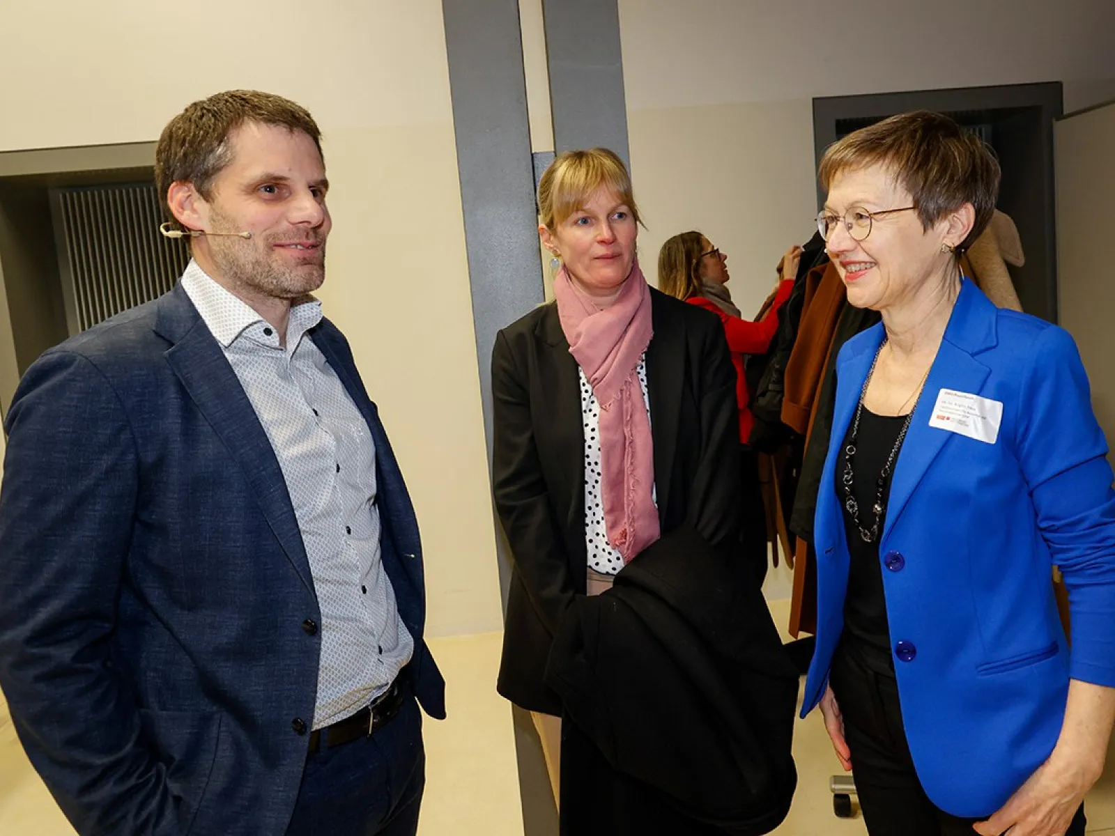 Drei Personen unterhalten sich bei einer Veranstaltung an der Universität Liechtenstein. Eine Frau trägt einen blauen Blazer, eine andere einen schwarzen Mantel mit rosa Schal, und der Mann trägt ein dunkelblaues Sakko.