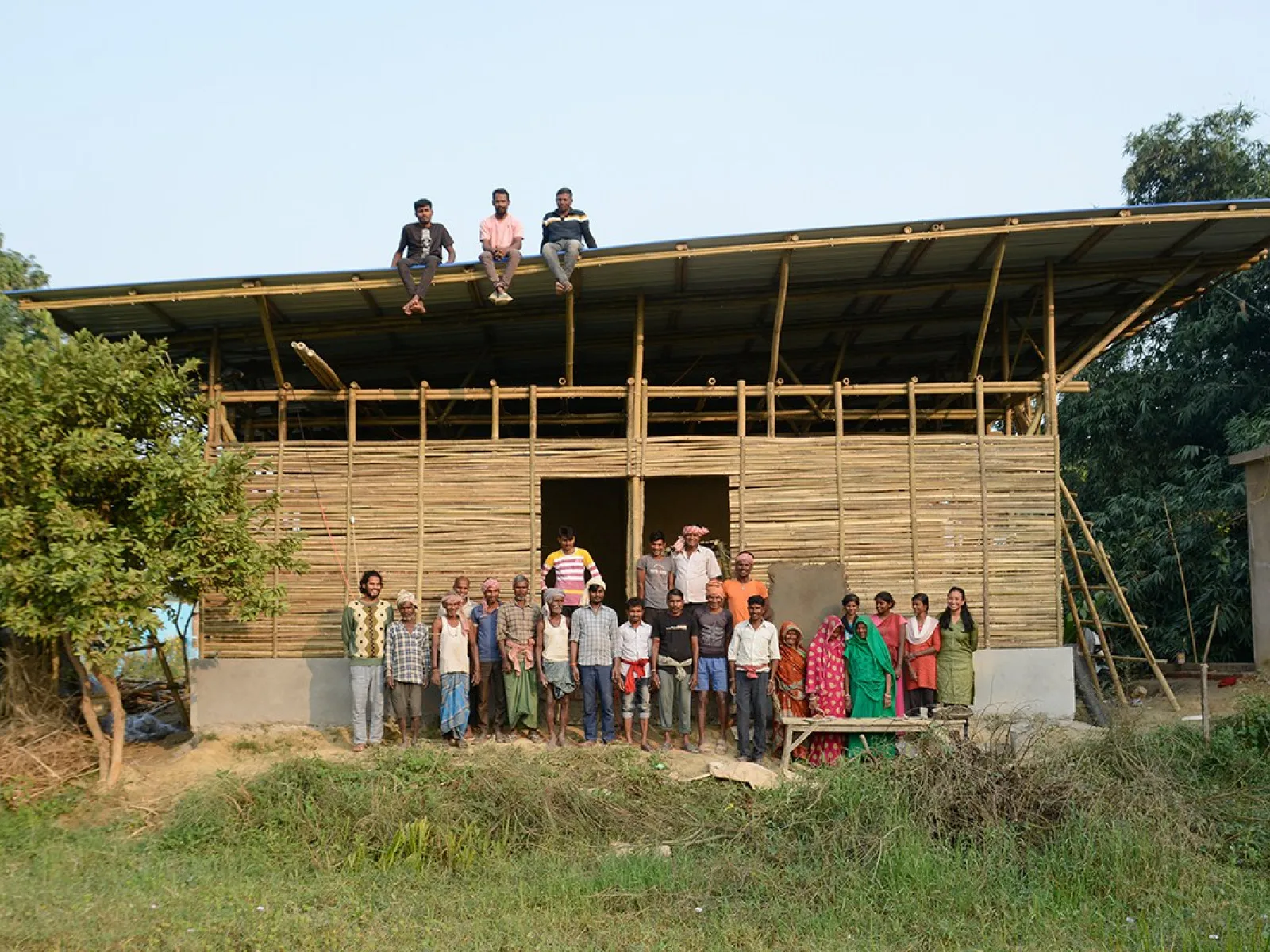 Eine Gruppe von Menschen steht vor einem im Bau befindlichen Gebäude aus Bambus in Bahuarwa, Indien. Drei Personen sitzen auf dem Dach. Das Bild zeigt den Fortschritt eines Schulbauprojekts, das von der Universität Liechtenstein unterstützt wird.