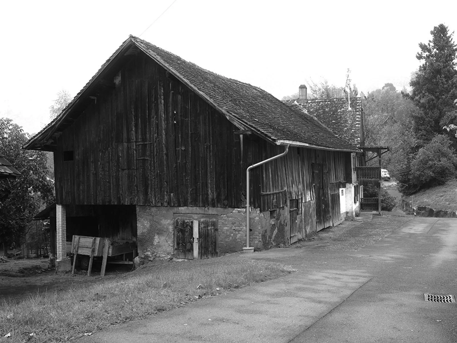 Altes Strickbau-Gebäude an der Kapfstrasse in Eschen, Liechtenstein, mit Holzfassade und schrägem Dach, umgeben von Bäumen und einem asphaltierten Weg.