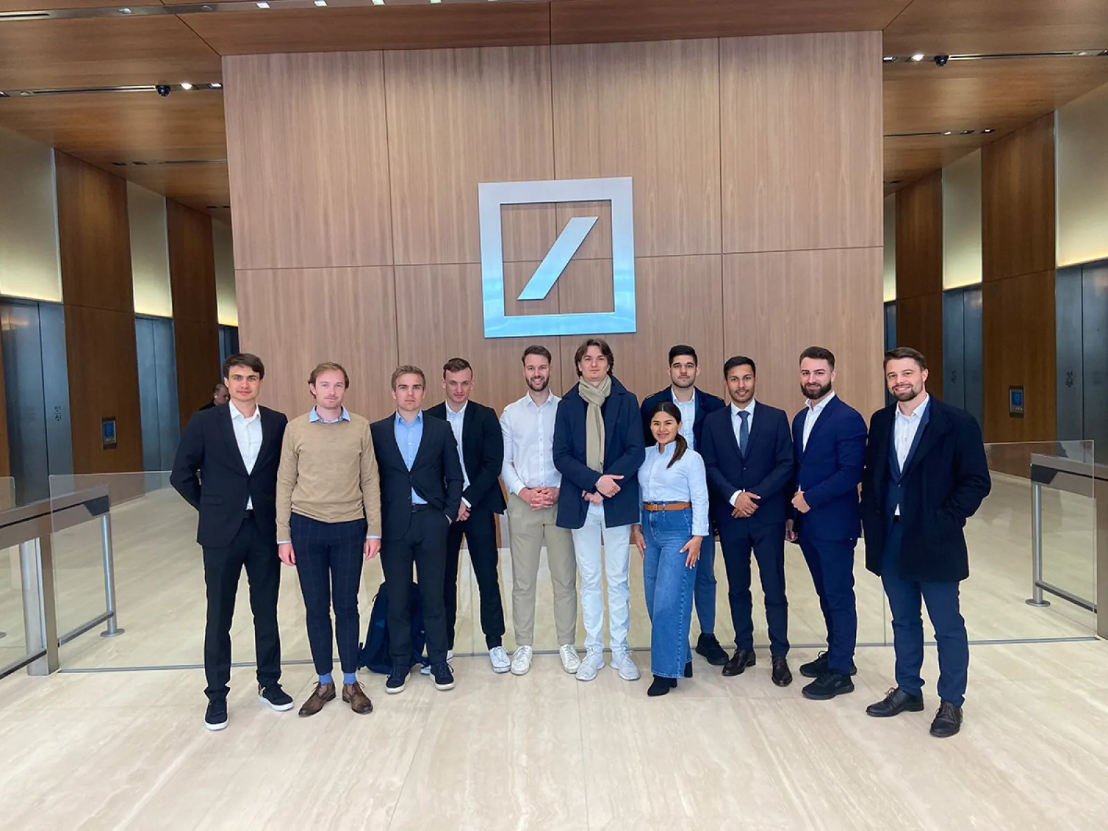 Eine Gruppe von Studierenden und Begleitpersonen steht in einem modernen Foyer vor dem Logo einer bekannten Bank. Sie tragen formelle Kleidung und lächeln in die Kamera.