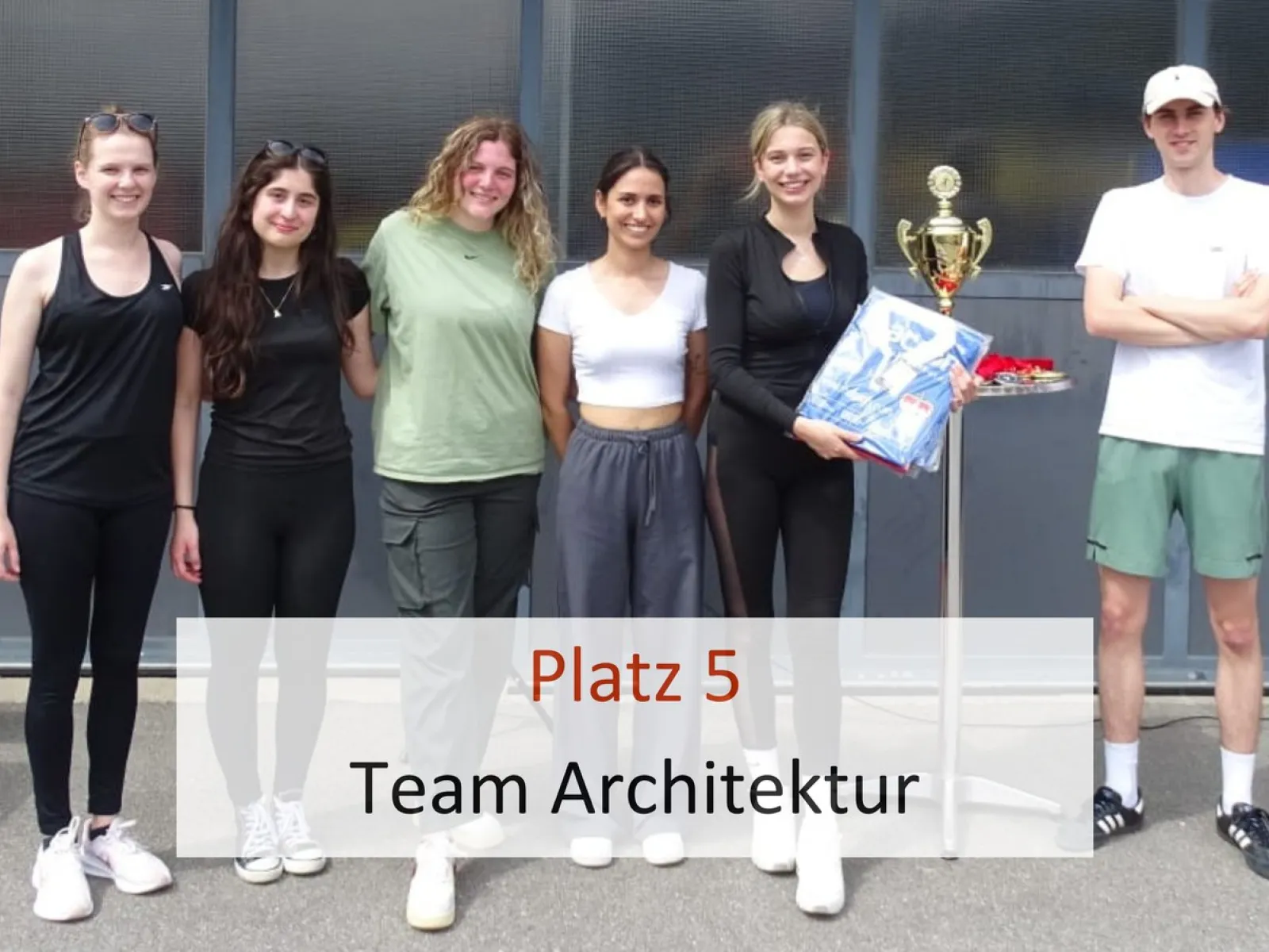 Sechs Personen stehen nebeneinander und lächeln in die Kamera. Eine Person hält einen Pokal und eine Schachtel. Im Vordergrund steht der Text "Platz 5 Team Architektur".