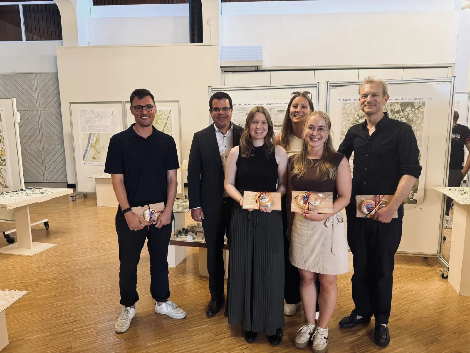 Gruppe von Studierenden und Dozenten der Universität Liechtenstein präsentiert Architekturprojekte im Rahmen eines Advanced Studios. Im Hintergrund sind Plakate und Modelle zu sehen, die ortsbauliche Strategien für die Gemeinde Ruggell zeigen.