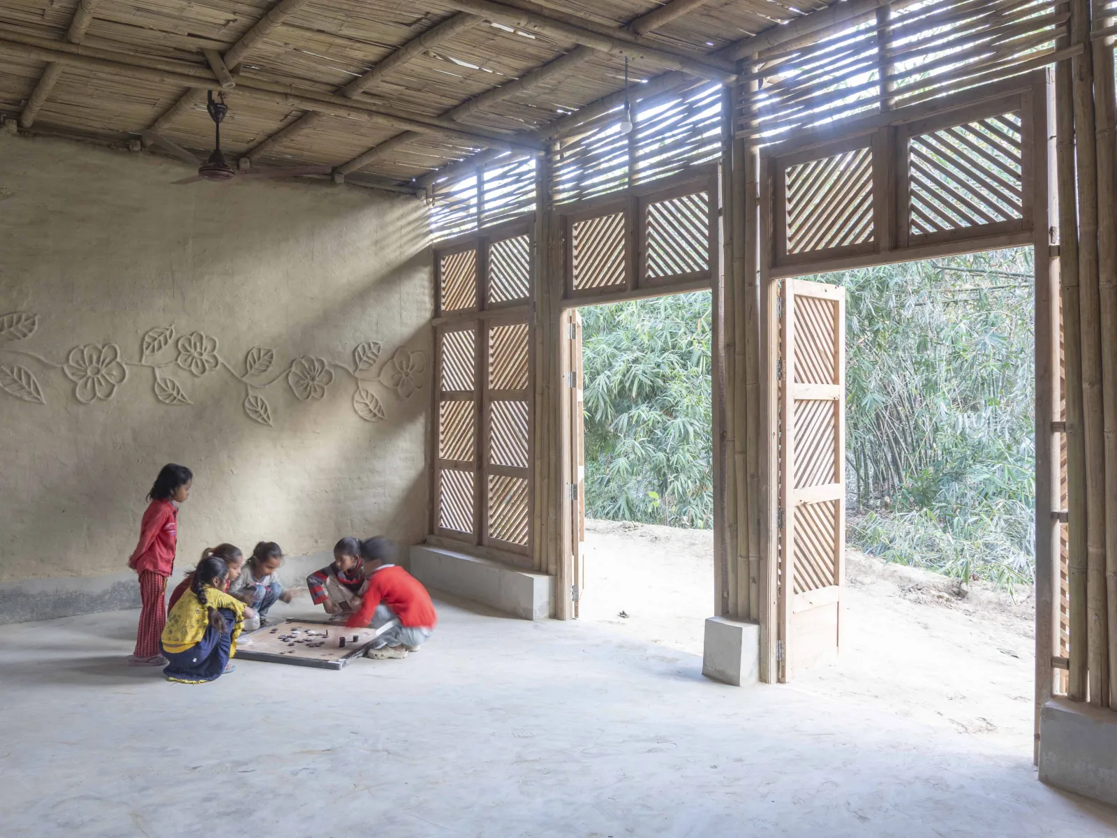 Kinder spielen auf dem Boden eines offenen, aus Naturmaterialien gebauten Raums mit Blick ins Grüne.
