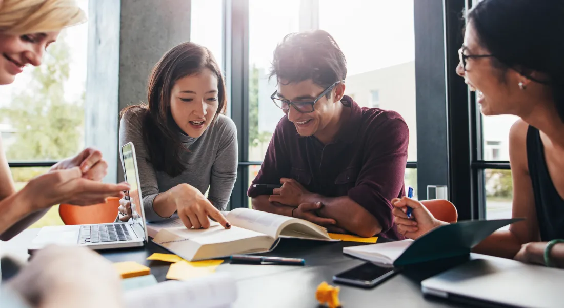 Eine Gruppe arbeitet gemeinsam an einer Aufgabe, während sie an einem Tisch sitzt, auf dem Laptops, Bücher und Notizen verteilt sind. Die Atmosphäre ist dabei entspannt und freundlich.