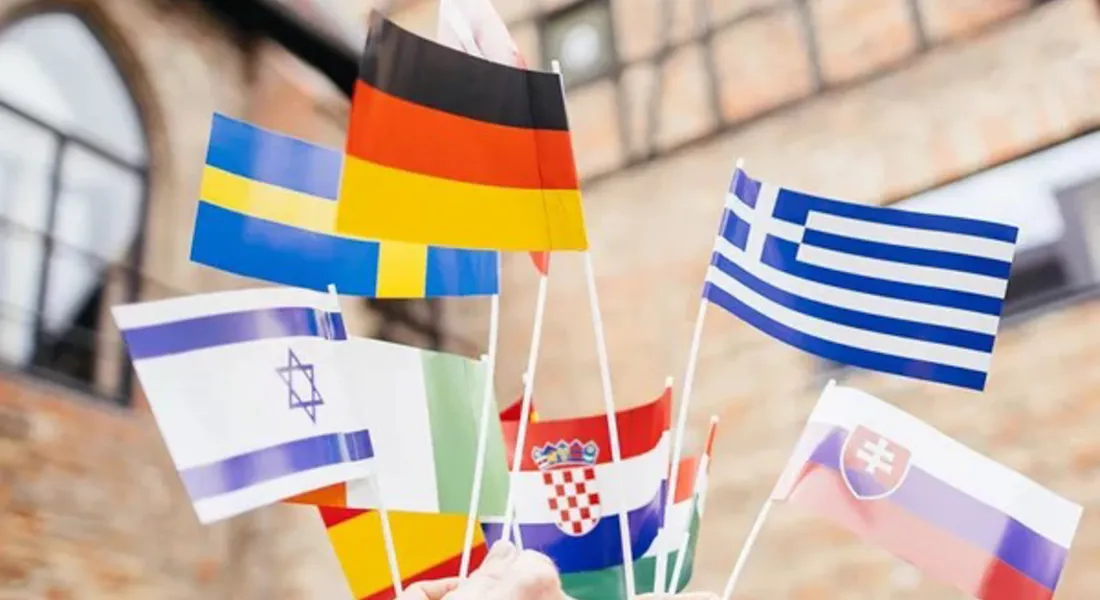 Mehrere Hände halten kleine internationale Flaggen in die Höhe, darunter unter anderem die Flaggen von Deutschland, Griechenland, Schweden, Israel, Kroatien, der Slowakei, Italien und Litauen. Im Hintergrund ist ein historisches Gebäude zu sehen. Das Bild symbolisiert internationale Vielfalt und kulturellen Austausch an der Universität Liechtenstein.