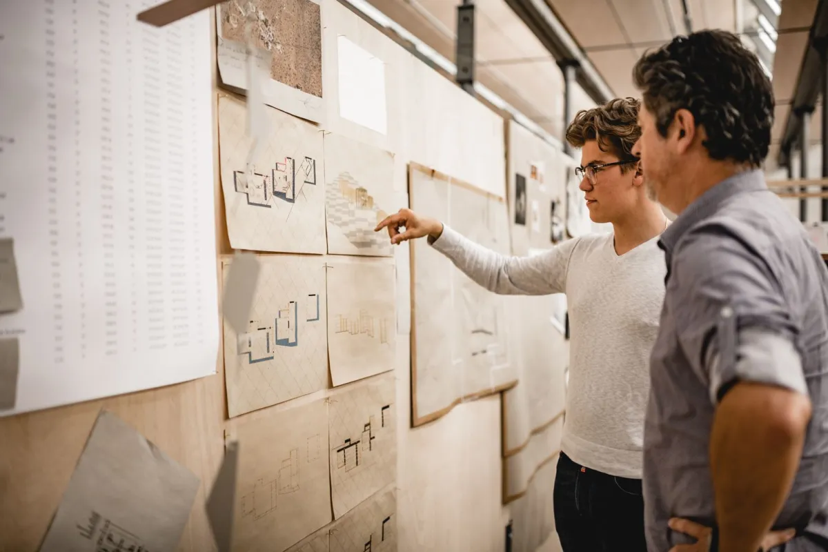 Studierender und Dozent sprechen über Architekturpläne die an einer Pinnwand hängen