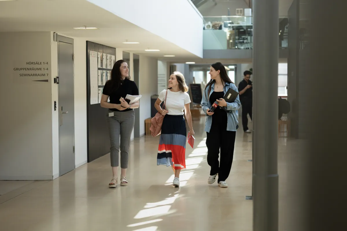 Drei Studentinnen gehen lächelnd durch das lichtdurchflutete Foyer mit Unterlagen in der Hand.