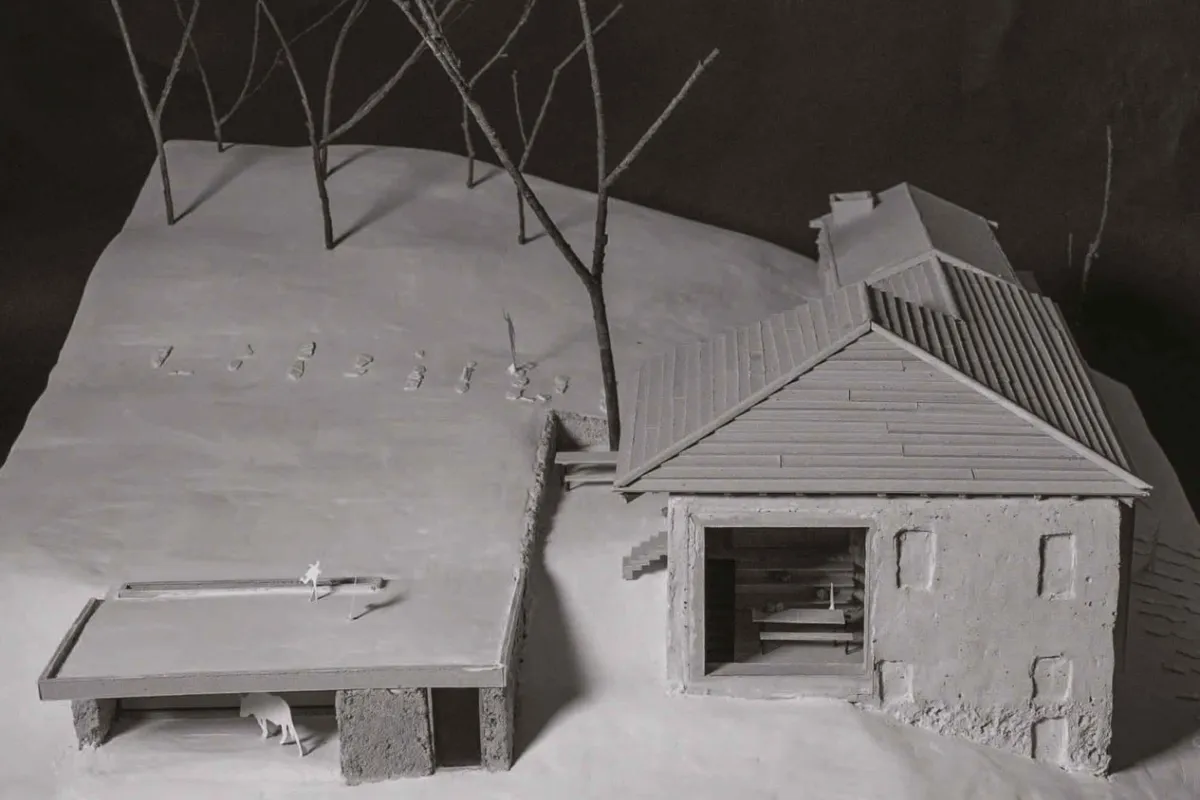 Ein Architekturmodell zeigt ein Gebäude mit Satteldach und angrenzendem offenen Unterstand auf einer schneebedeckten Landschaft. Im Hintergrund stehen stilisierte Bäume.