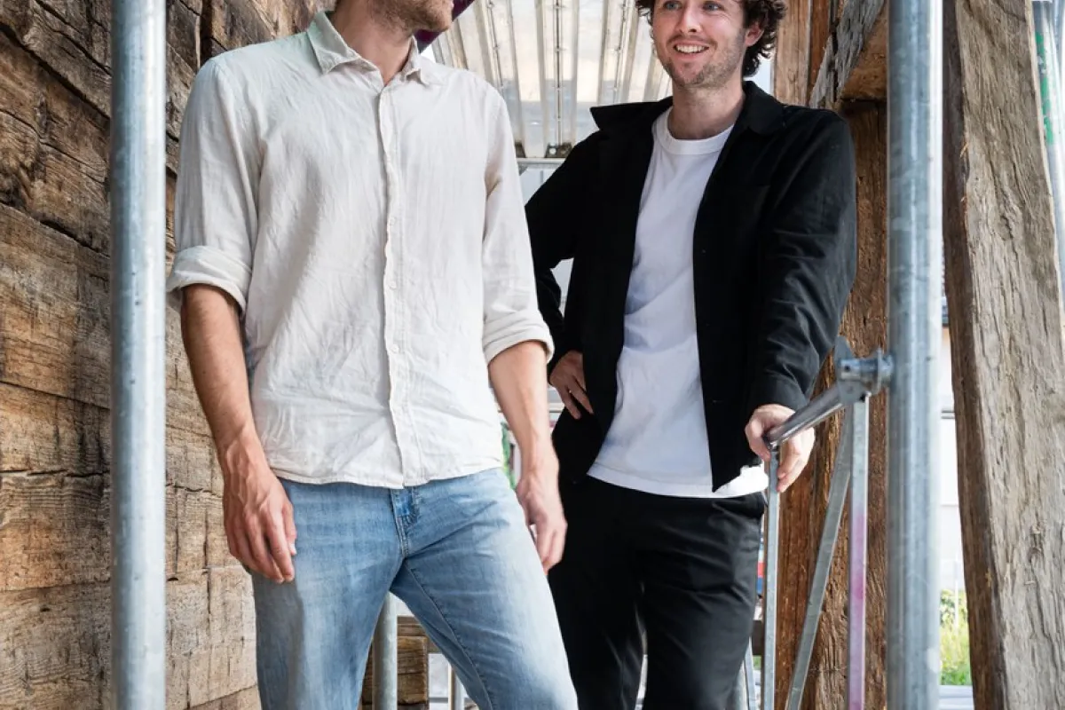 Zwei junge Männer stehen lächelnd auf einem Gerüst in einem Holzbau. Sie tragen legere Kleidung und wirken entspannt. Die Umgebung deutet auf eine Baustelle oder ein architektonisches Projekt hin.