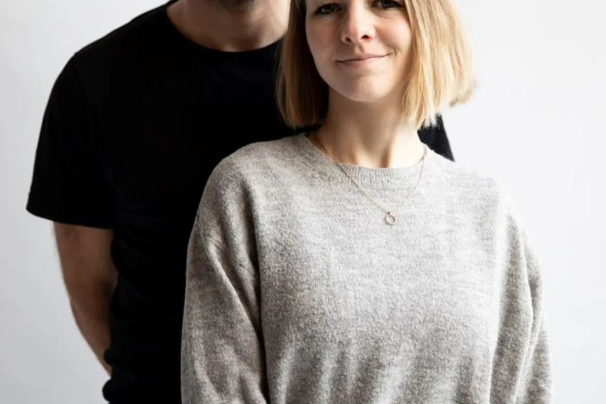 Ein Mann und eine Frau stehen vor einem neutralen Hintergrund. Beide blicken direkt in die Kamera. Die Frau trägt einen grauen Pullover, der Mann ein schwarzes T-Shirt.
