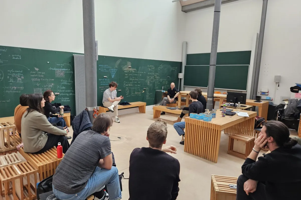 Eine Gruppe von Studierenden sitzt in einem modernen Seminarraum der Universität Liechtenstein, umgeben von Tafeln mit Notizen und Skizzen. Sie diskutieren angeregt, während ein Dozent vorne sitzt und zuhört.